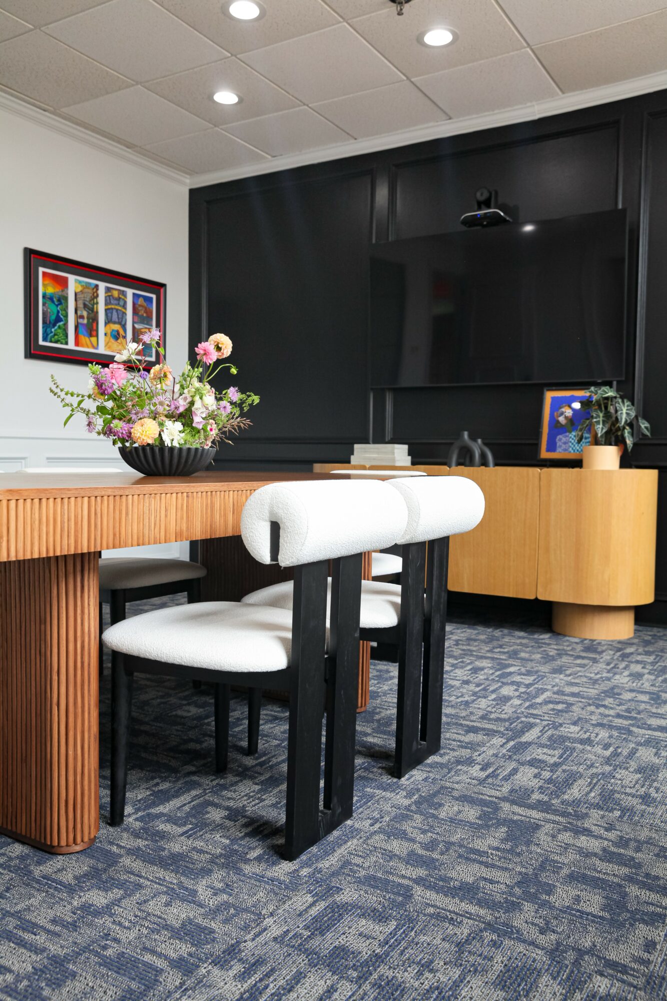 Room with a wooden table, white cushioned chairs, a black wall with a large TV, and floral arrangements.