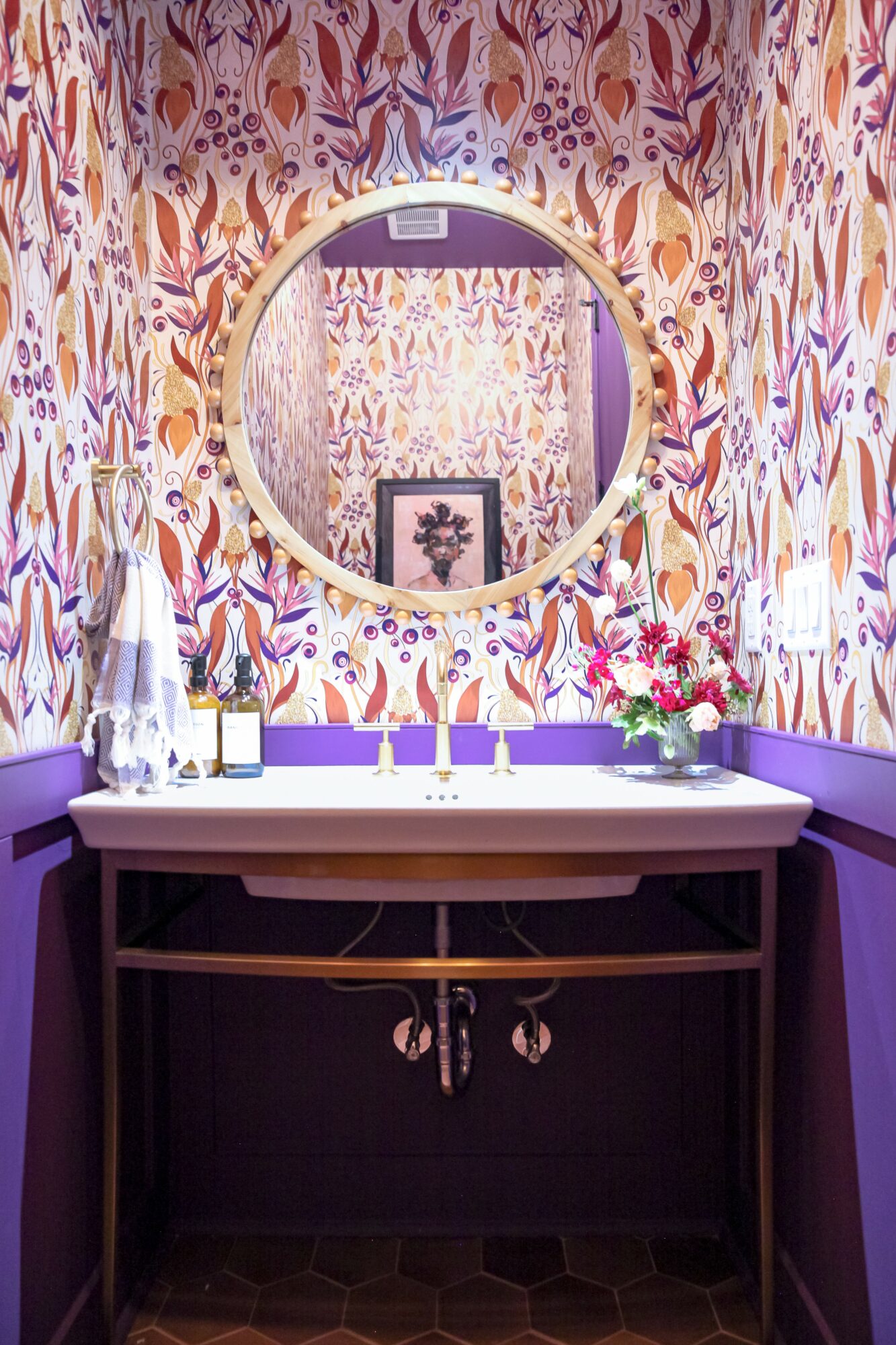 Bathroom vanity with a round mirror, floral wallpaper, purple walls, and a white sink with flowers and toiletries.