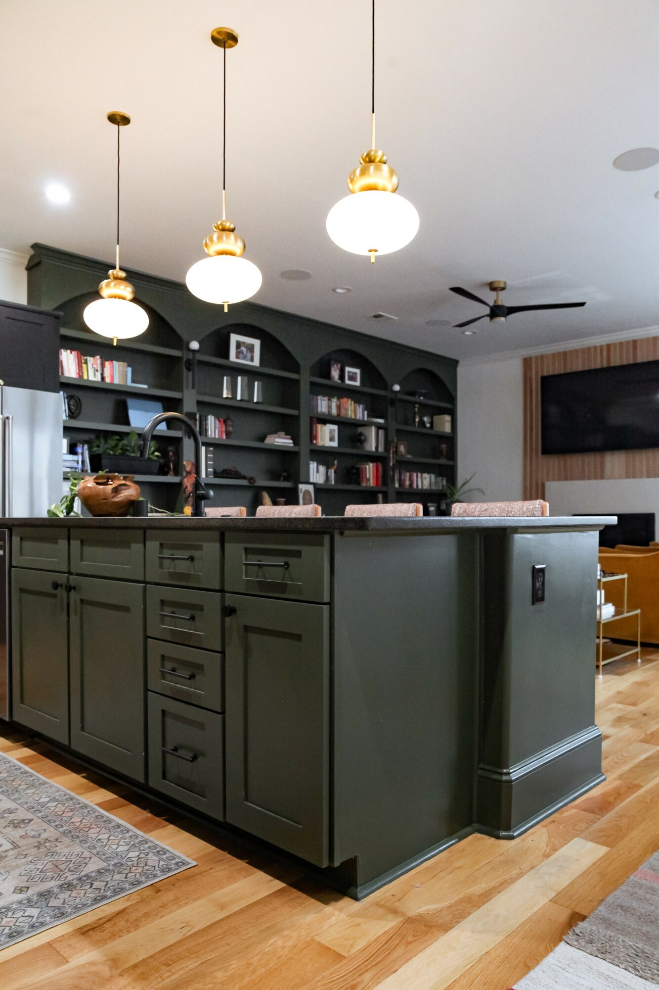 Kitchen island with dark green cabinets, pendant lights, and a bookshelf wall in the background, hardwood floor, and ceiling fan.