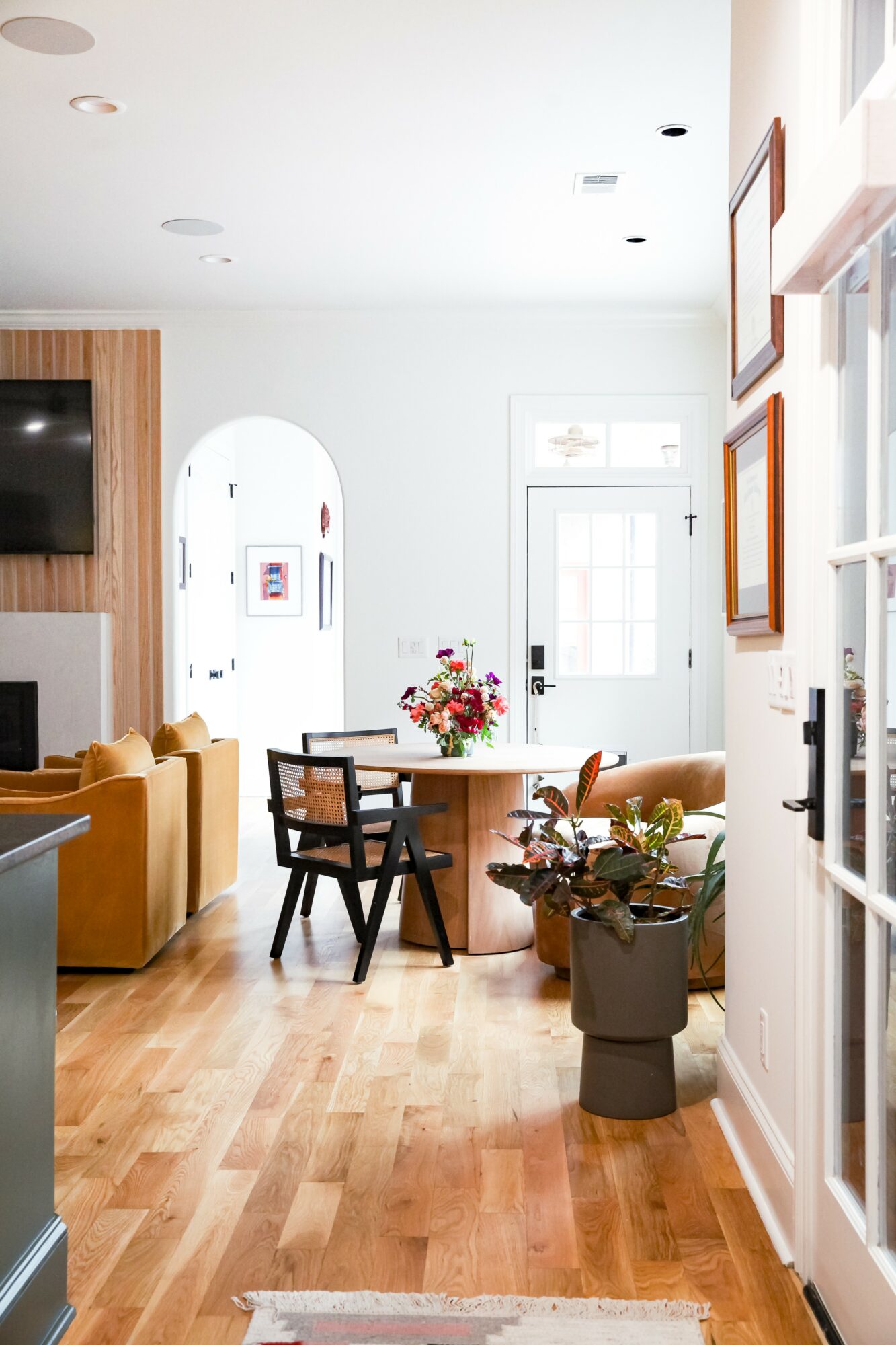 Living room with dining table, chairs, sofa, and wall decorations, bright natural light, wooden flooring, and a door in background.