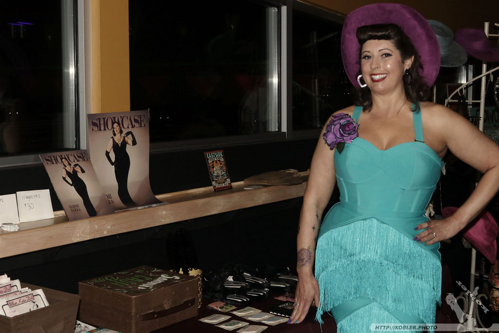 Woman in turquoise dress and purple hat smiling, standing indoors near a window with posters and items on a counter.