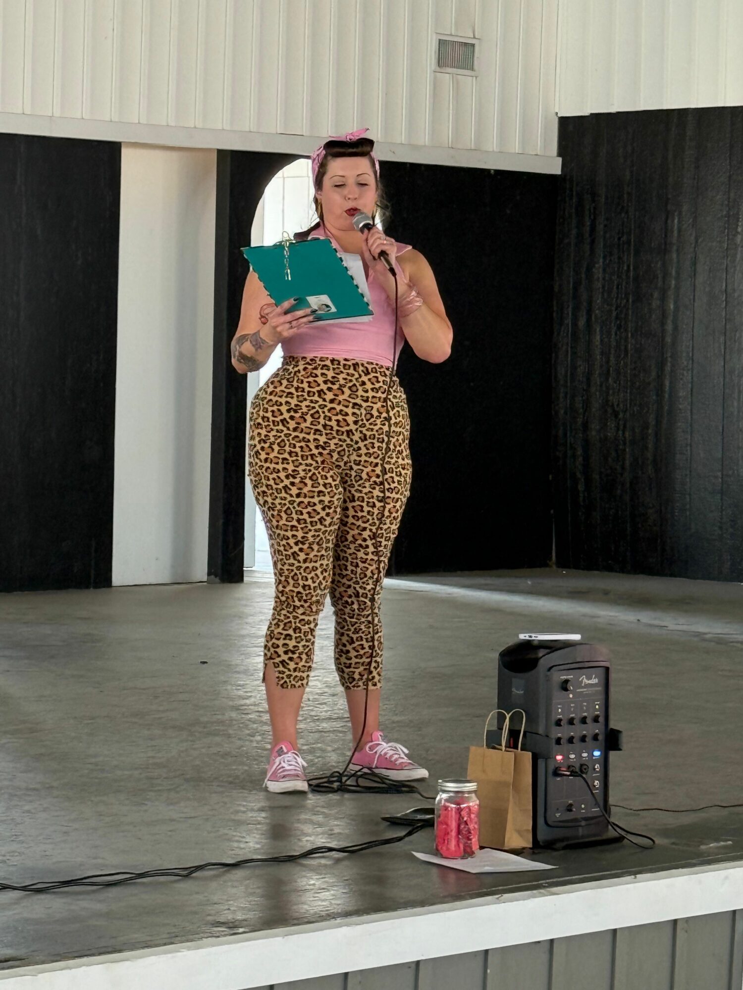 Person standing on stage holding a book, wearing leopard print pants and pink shoes, with equipment nearby.