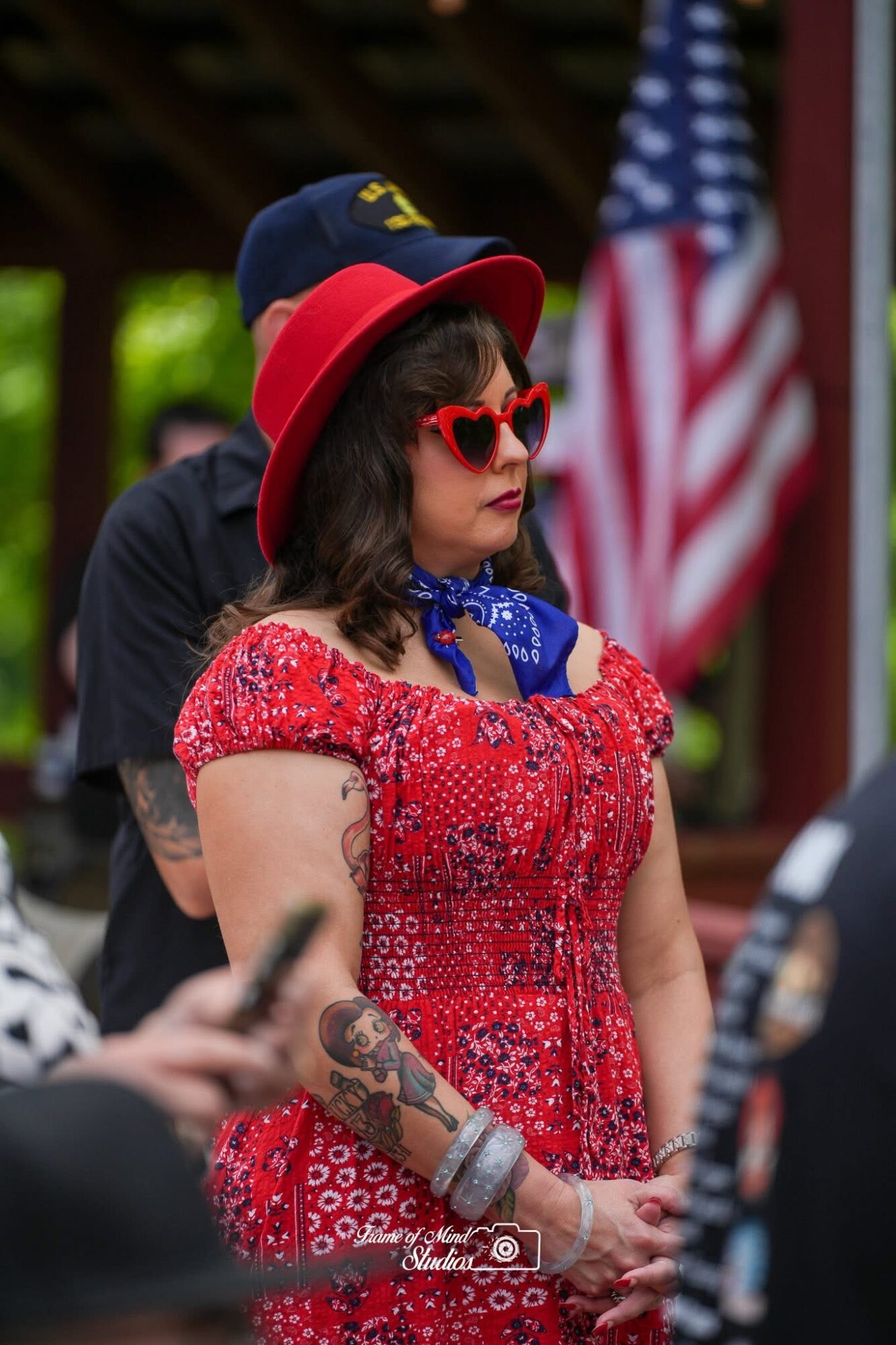 Woman wearing a red hat, red sunglasses, and a red patterned dress, standing outdoors with an American flag in the background.