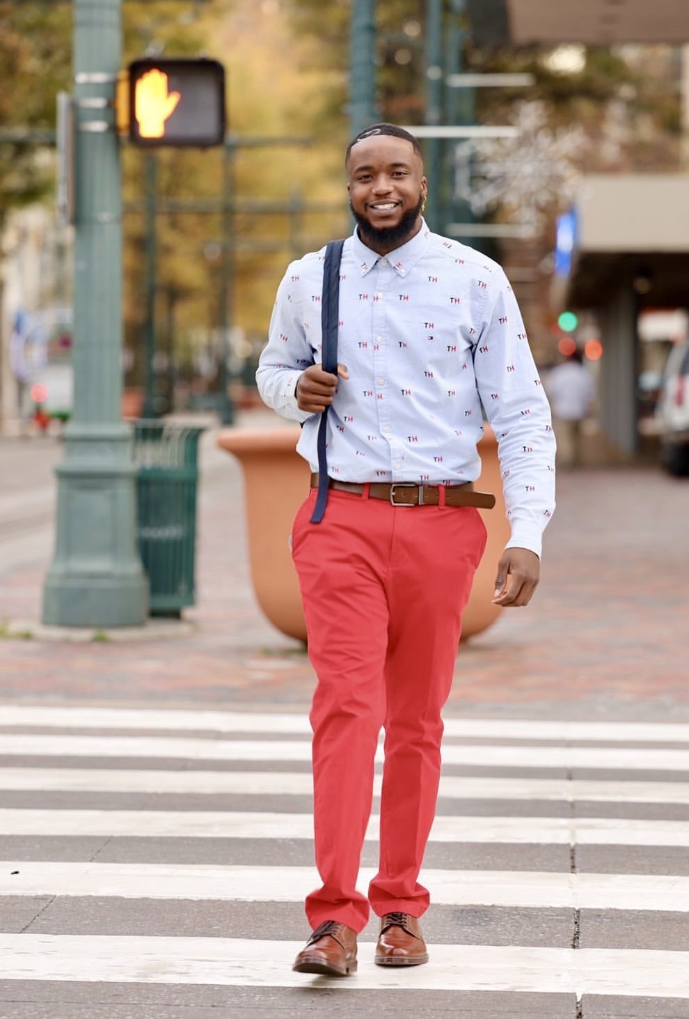 Man walking across a crosswalk holding a backpack, smiling, in an urban setting with trees and buildings.