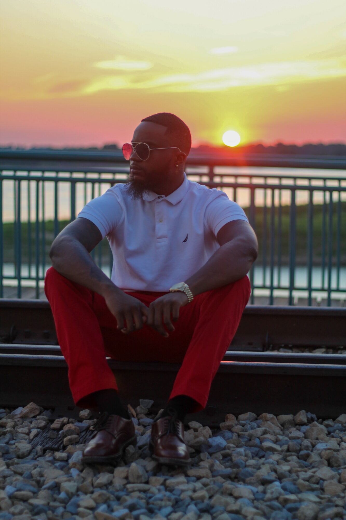 Person sitting outdoors near a railing during sunset, wearing sunglasses, a white shirt, and red pants.