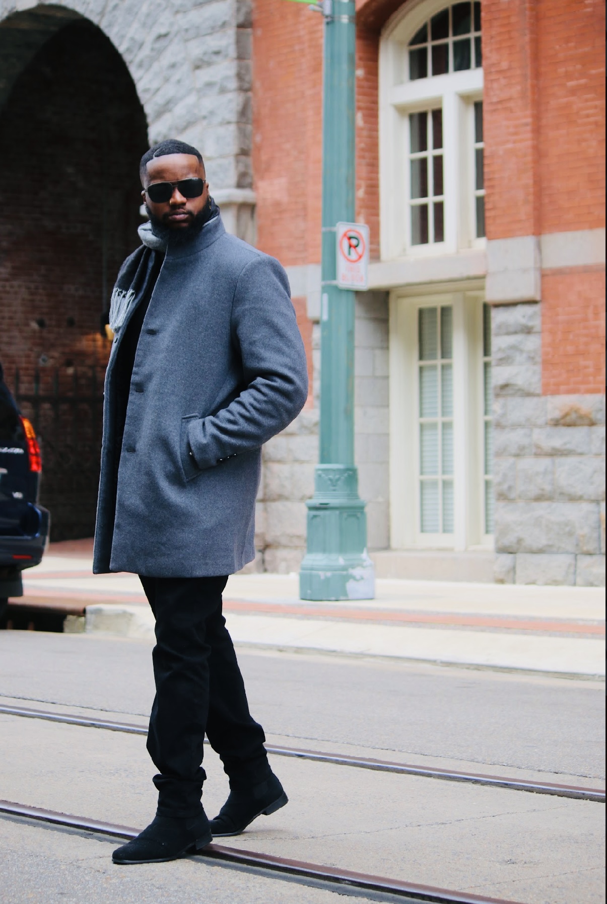 Man in gray coat and sunglasses walking on street with brick building and window behind him.