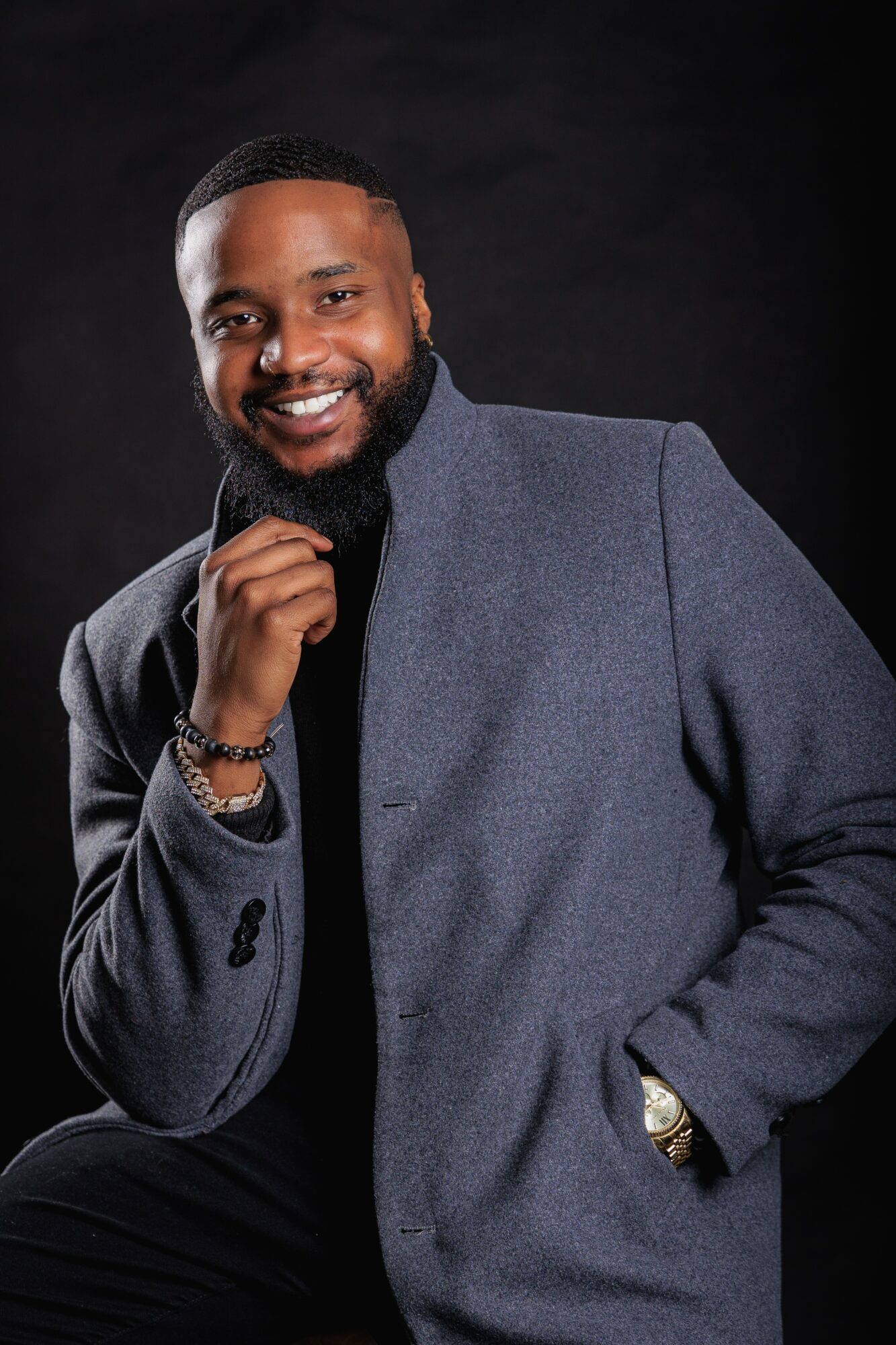 Man with a beard smiling, wearing a gray coat and black shirt, posing with hand near chin against black background.