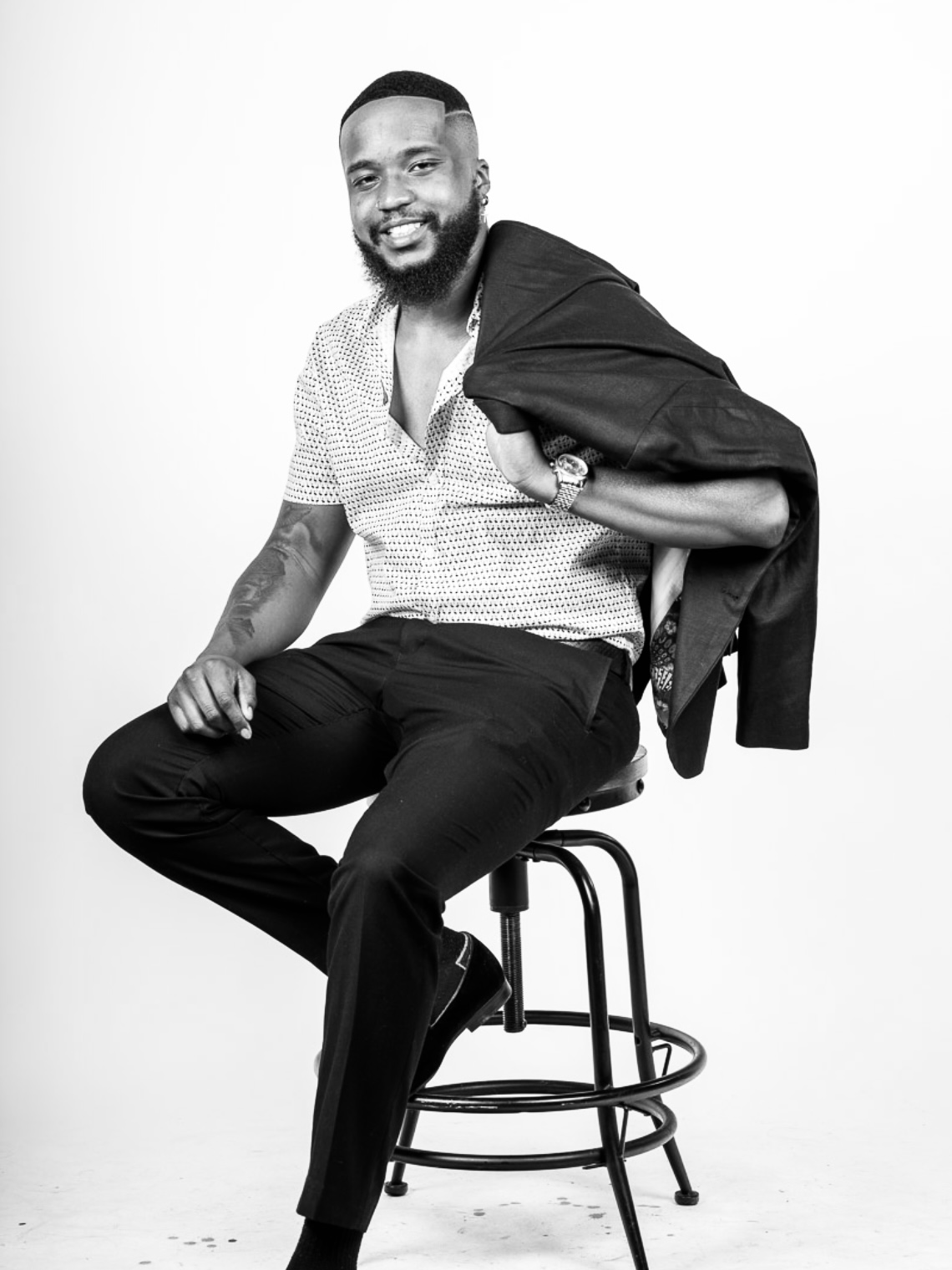 Man sitting on a stool, smiling, holding a jacket over his shoulder, wearing a short-sleeved shirt and dark pants.