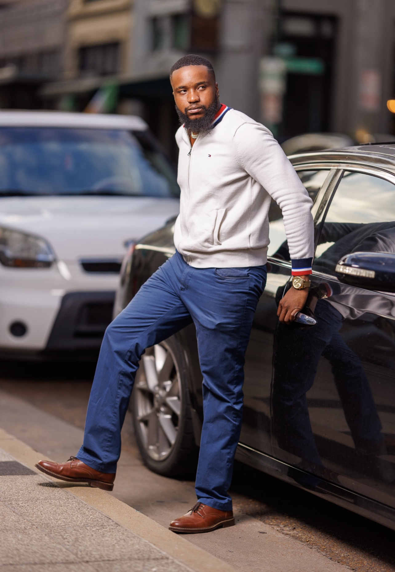 Man with beard in white jacket and blue pants leaning against a black car on city street, parked cars in background.