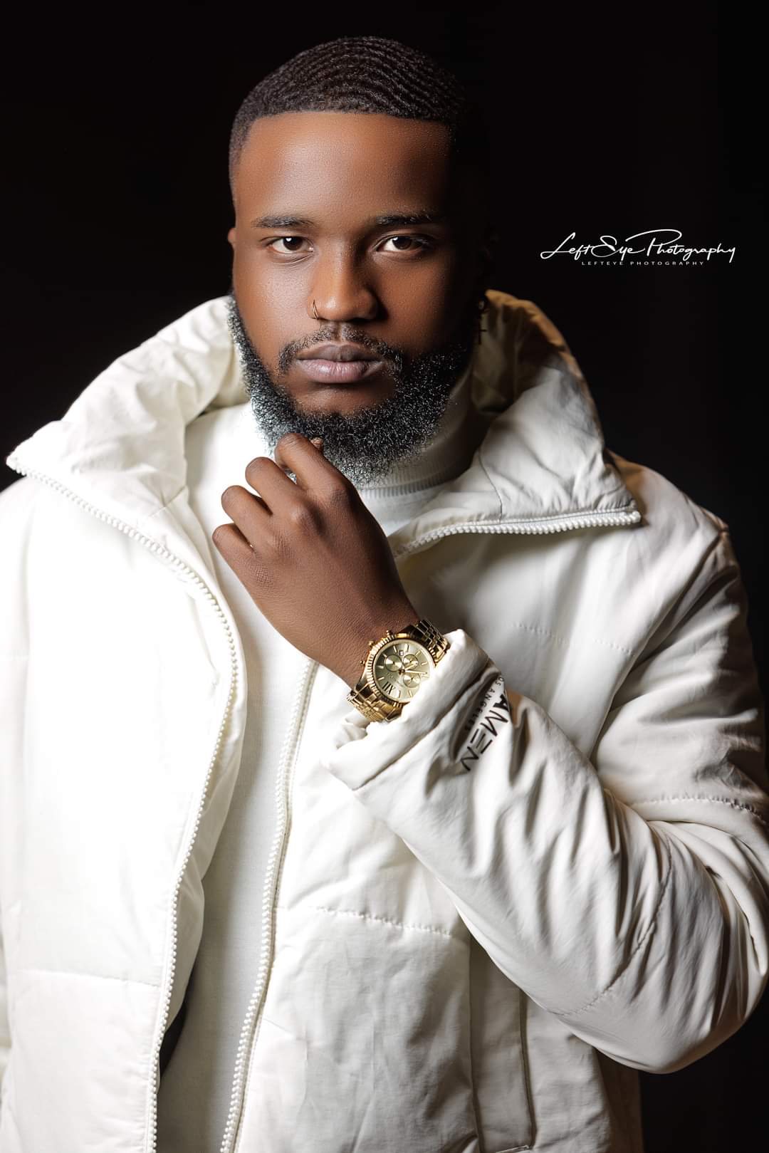Man with a beard wearing a white jacket and gold watch, looking at the camera against a dark background.