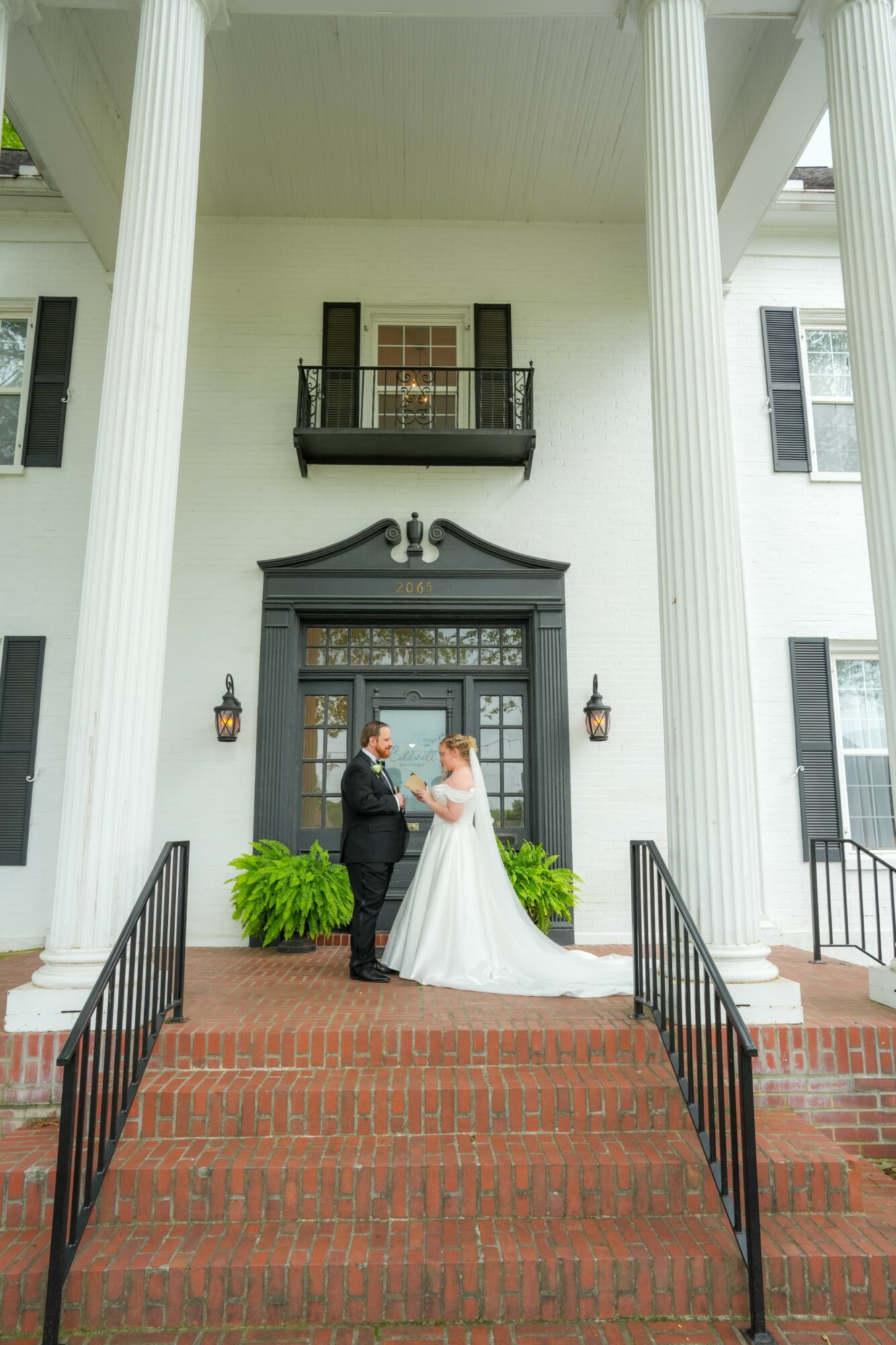 Bride and groom standing on steps in front of a large white house with tall columns, black door, and balcony above.