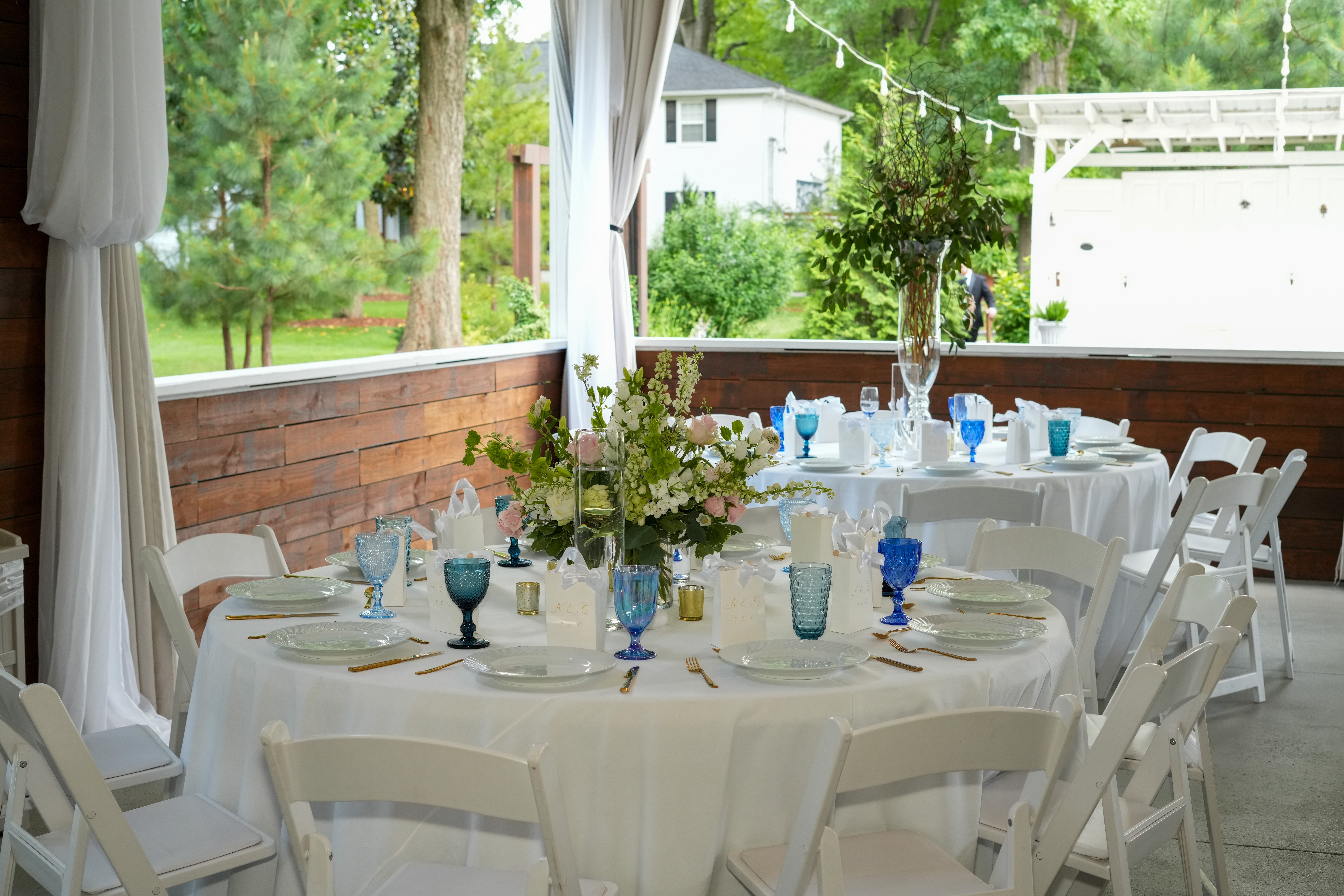Round table set with white tablecloth, floral centerpiece, blue glasses, and place settings, on a porch with trees outside.