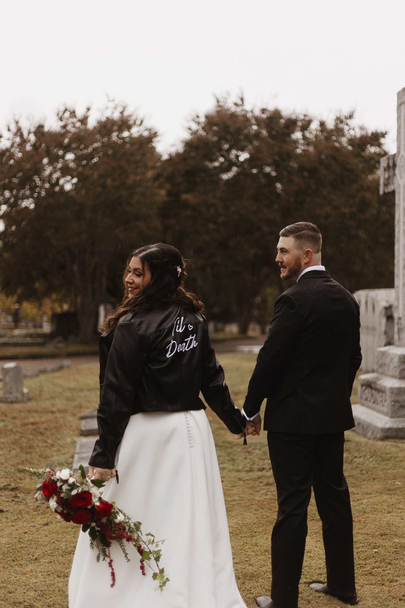 A couple holding hands outdoors, the woman holding a bouquet of flowers, trees in the background, overcast sky.
