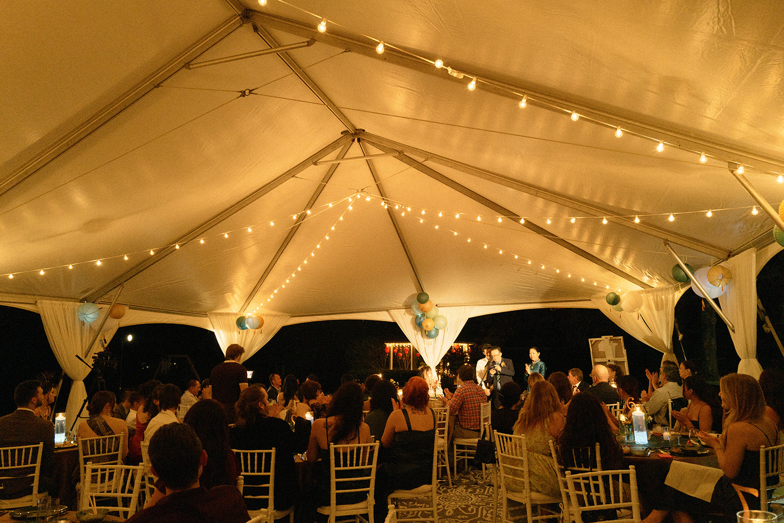 People seated at tables under a tent with string lights during an evening event.
