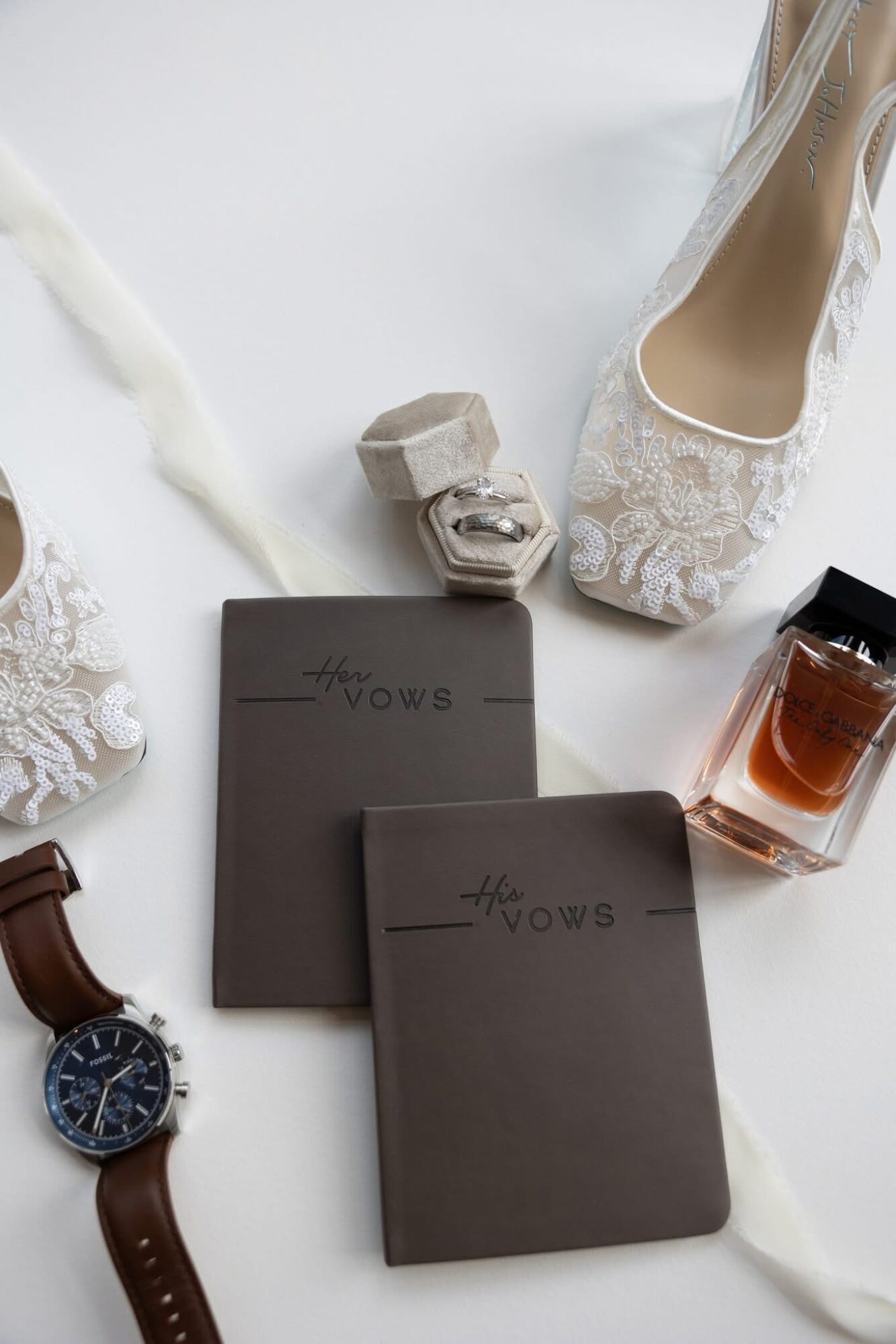 Wedding shoes, perfume, watch, and vow book on white surface with ribbon, arranged decoratively.