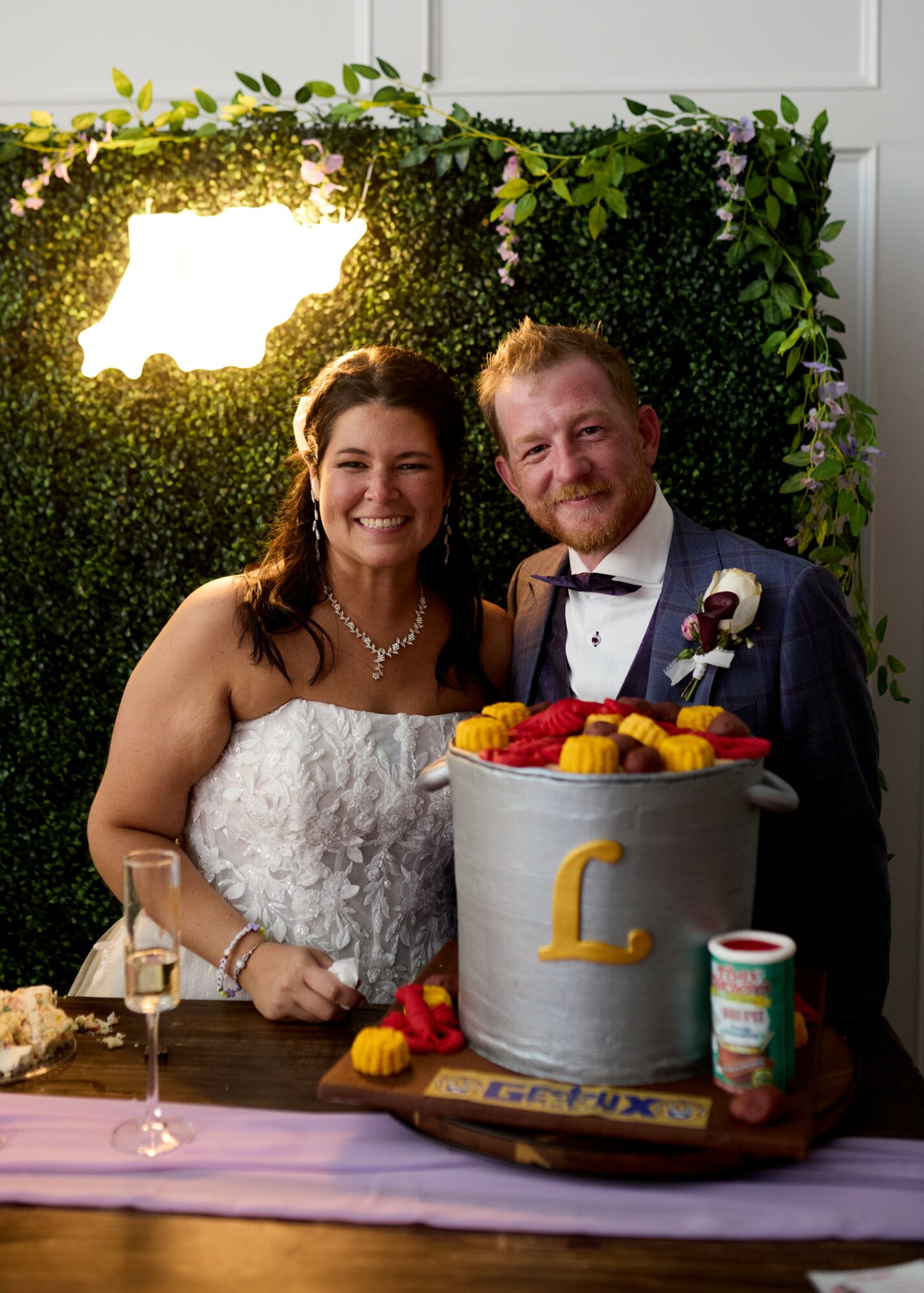 Smiling couple at celebration with cake, greenery backdrop, and table with drinks and decorations.