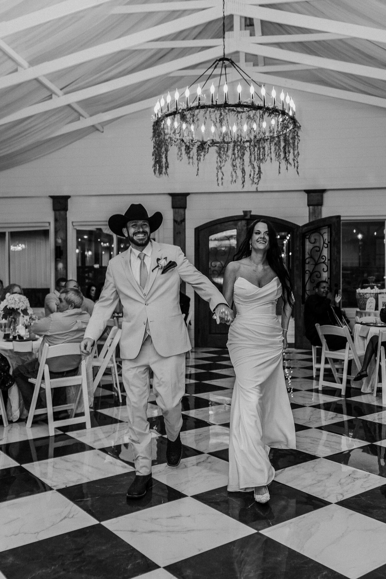 A couple walks hand in hand in a decorated event hall with a chandelier, checkered floor, and seated guests.