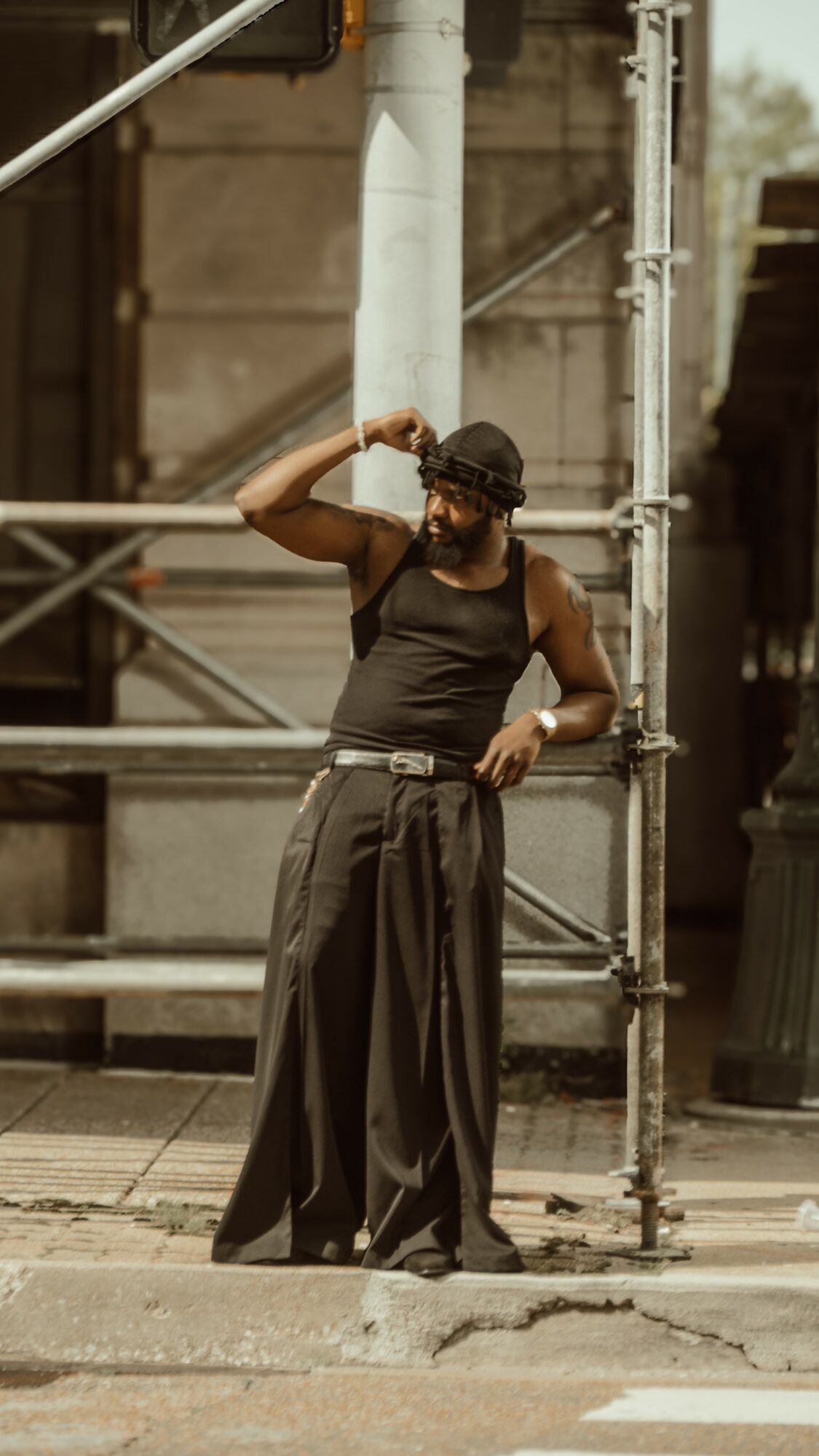 Person standing on sidewalk, wearing black tank top, wide pants, and sunglasses, adjusting their hat near scaffolding.