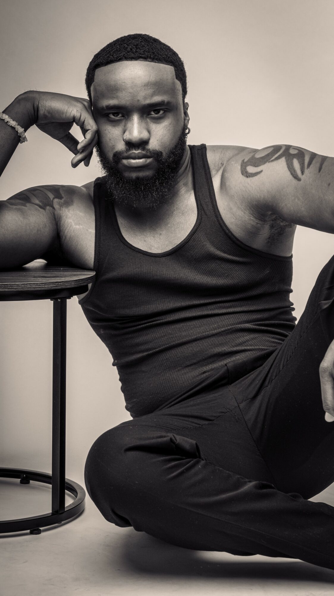 Man with a beard and tattoos sitting on the floor, resting his head on his hand, wearing a tank top.