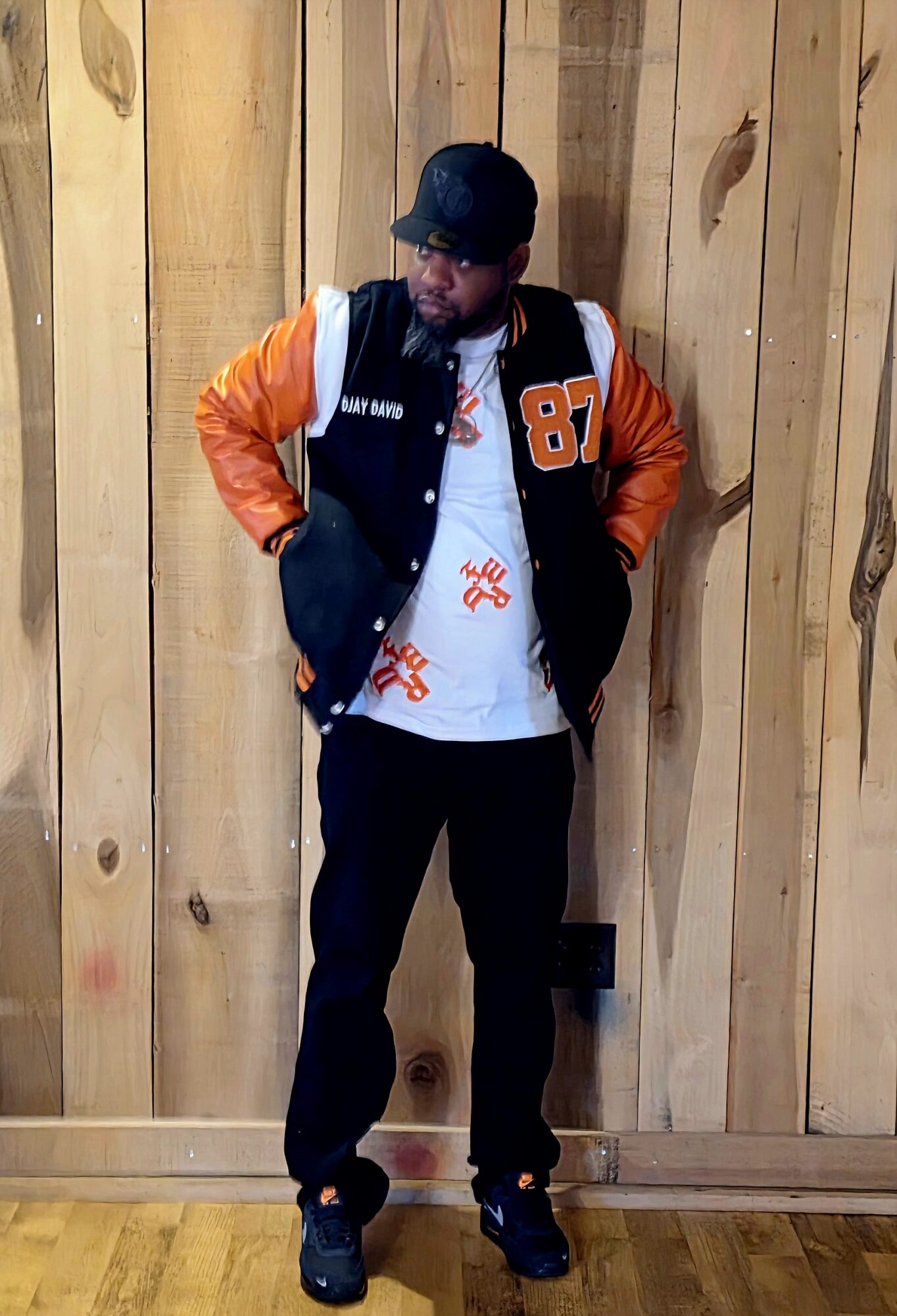 Person standing against wooden wall, wearing a black cap, orange and black jacket, white shirt, and black pants.