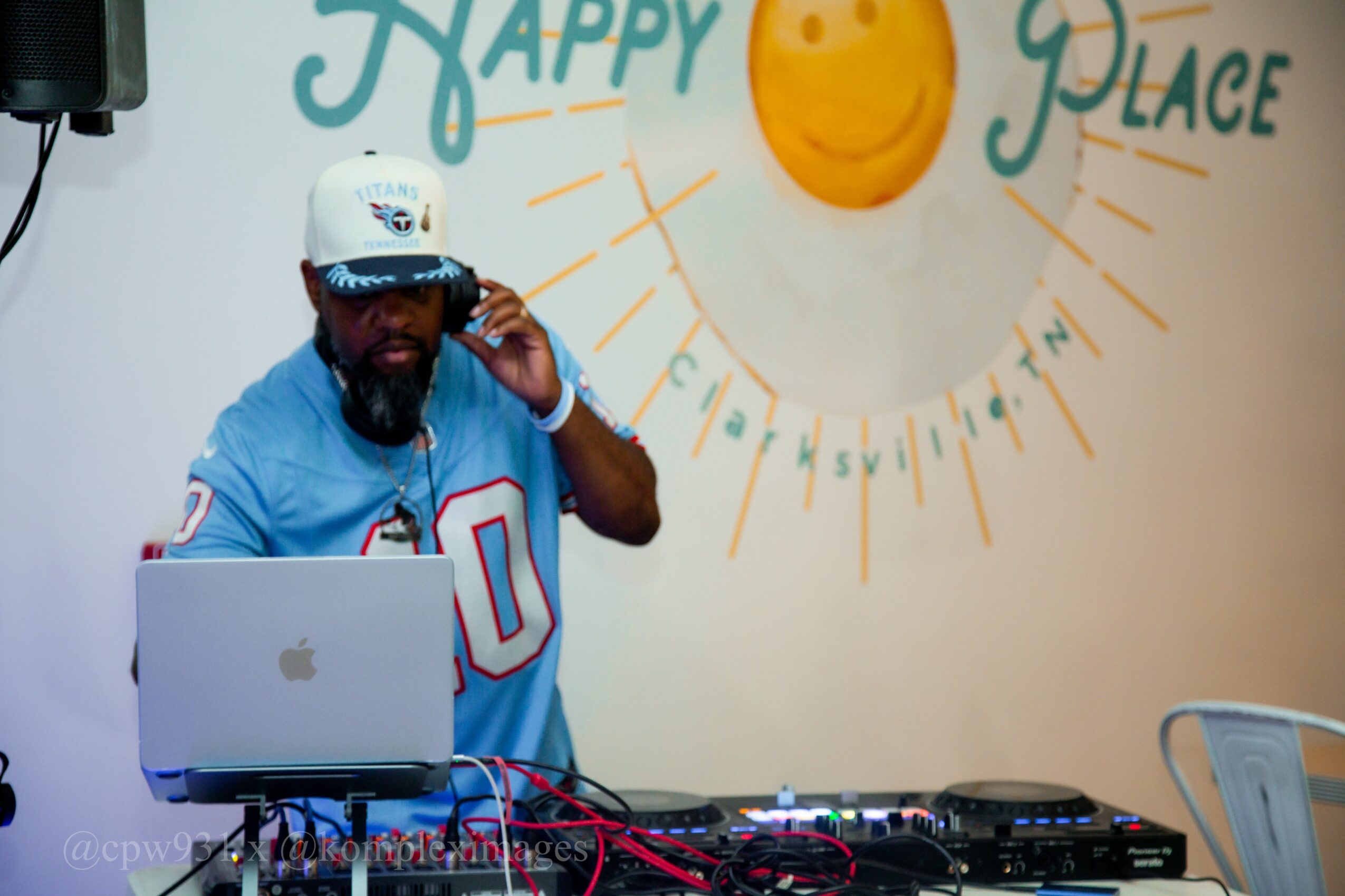 Man with a beard wearing a white cap and blue sports jersey, standing behind DJ equipment, talking on a phone, in front of a wall with a sun and the words 'Happy Place'.