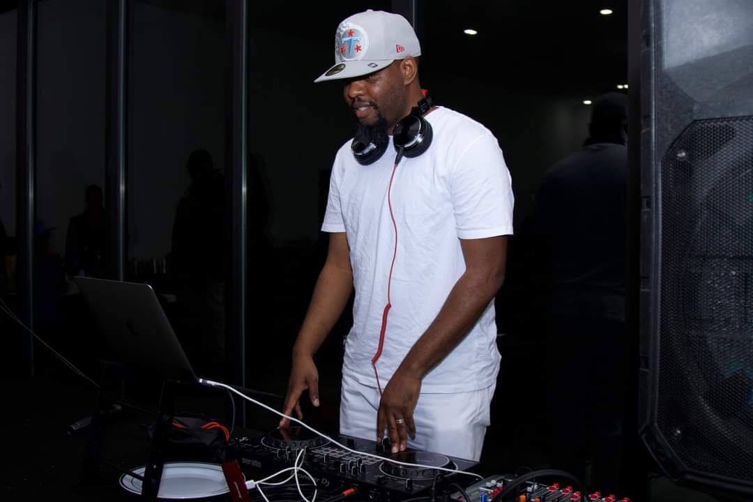 DJ wearing a white t-shirt, cap, and headphones, standing behind DJ equipment in a dark room.