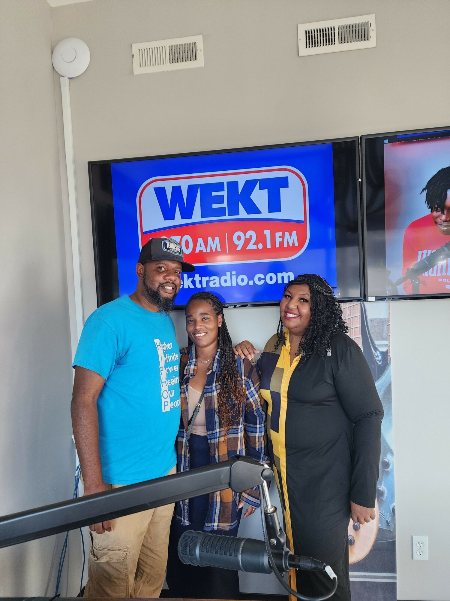 Two people standing in front of a TV screen displaying a radio station logo, one wearing a black blazer and the other in a blue shirt.