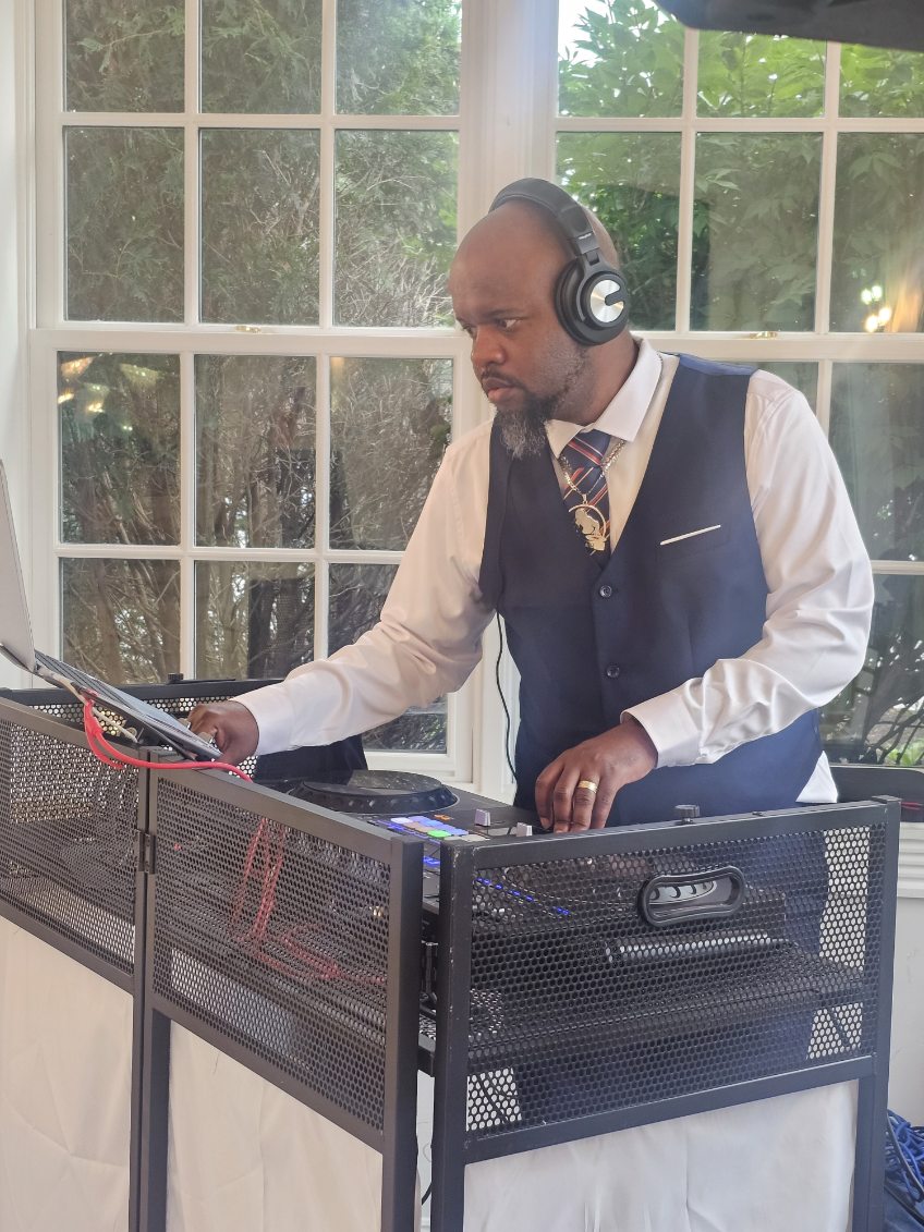 Man wearing headphones and formal attire, working with DJ equipment indoors near large window with greenery outside.