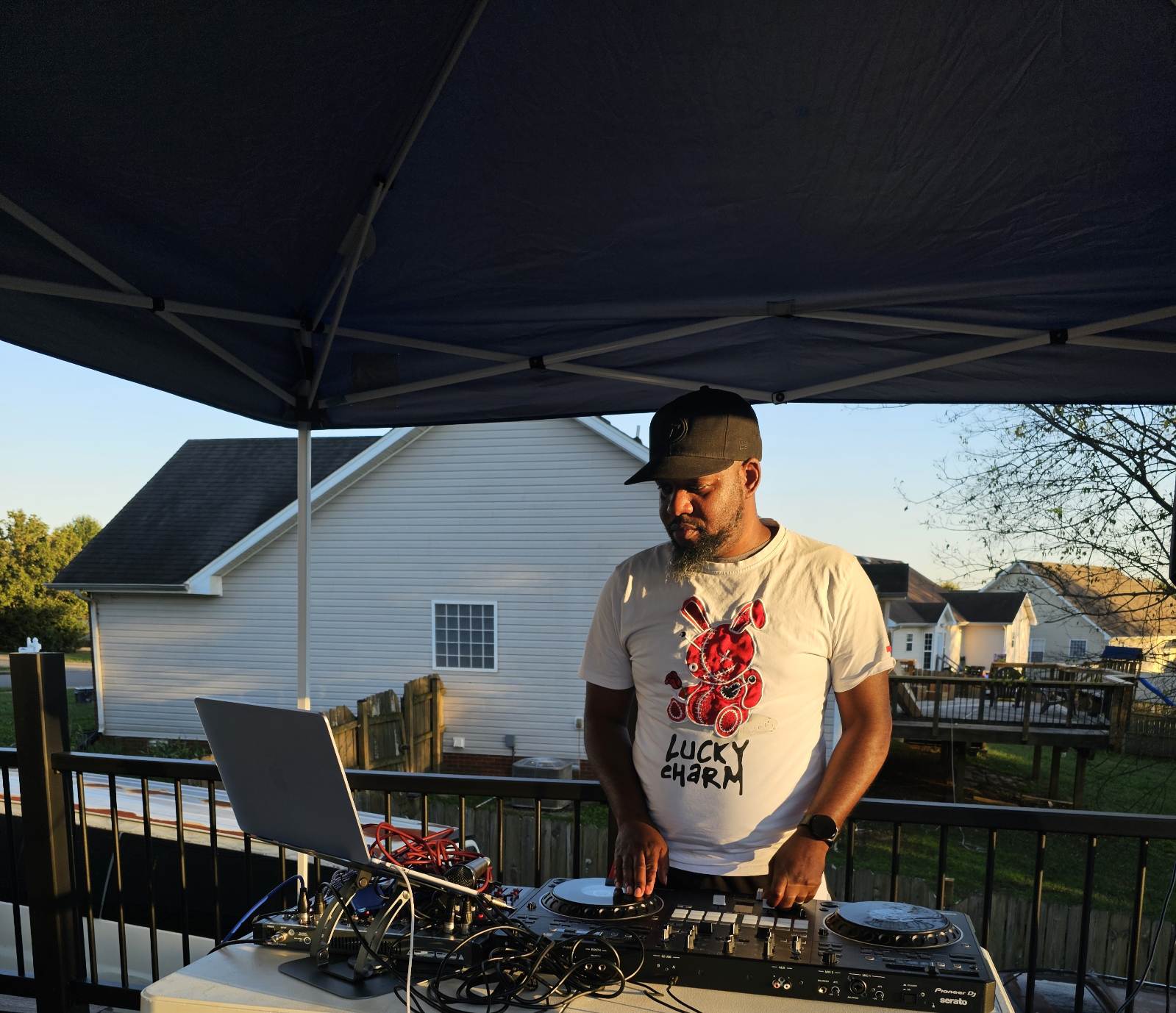 Man wearing a cap and sunglasses DJing outdoors under a canopy, with houses and trees in the background.