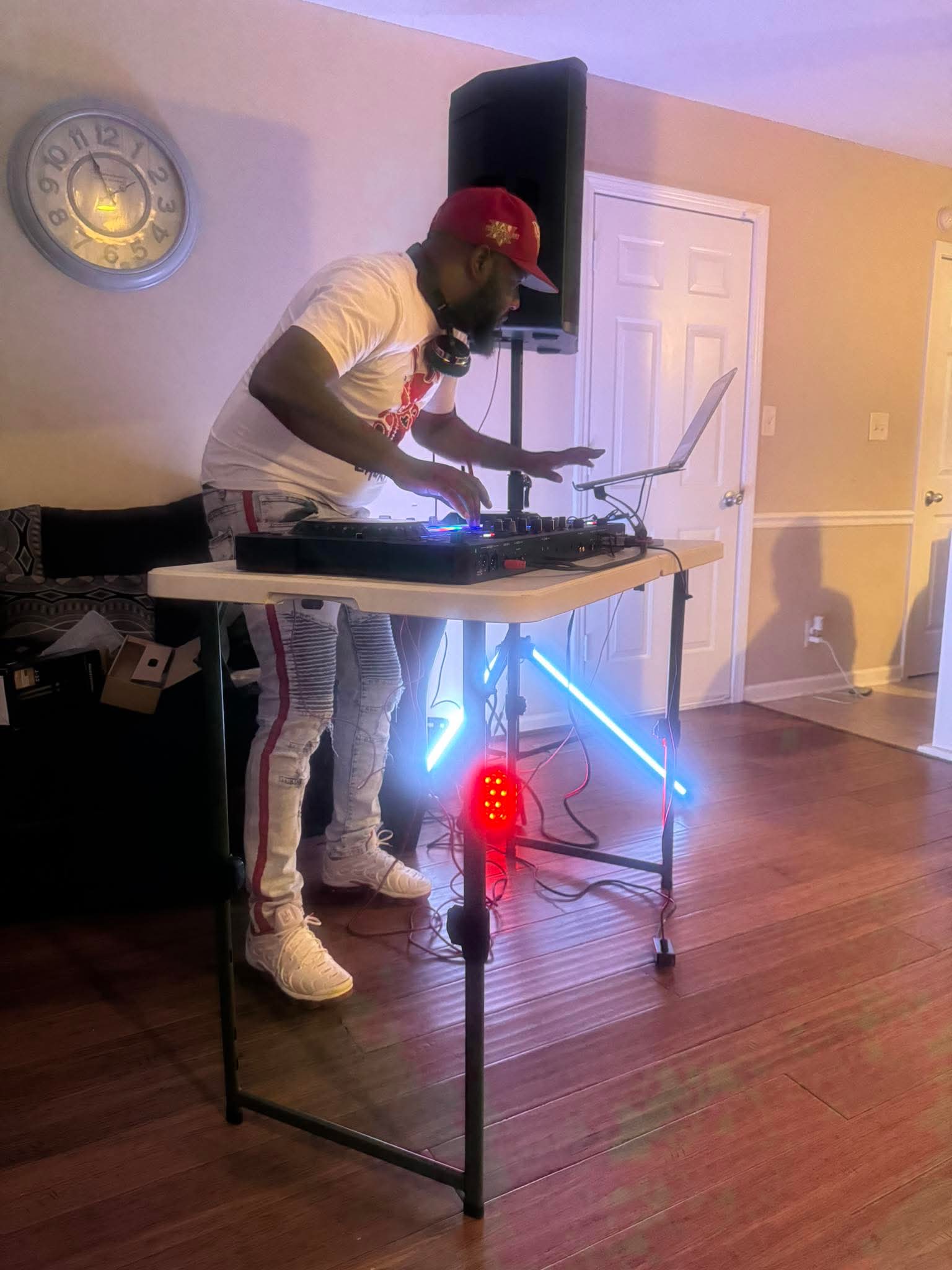 Person wearing a red cap and white shirt DJing with equipment on a table, in a room with hardwood floor and beige walls.