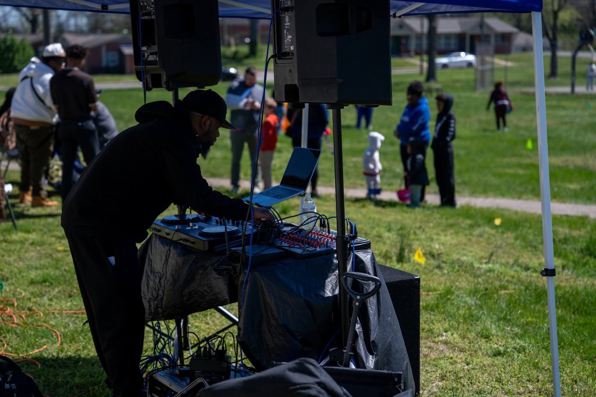 Person operating DJ equipment outdoors with people in the background on a grassy field.