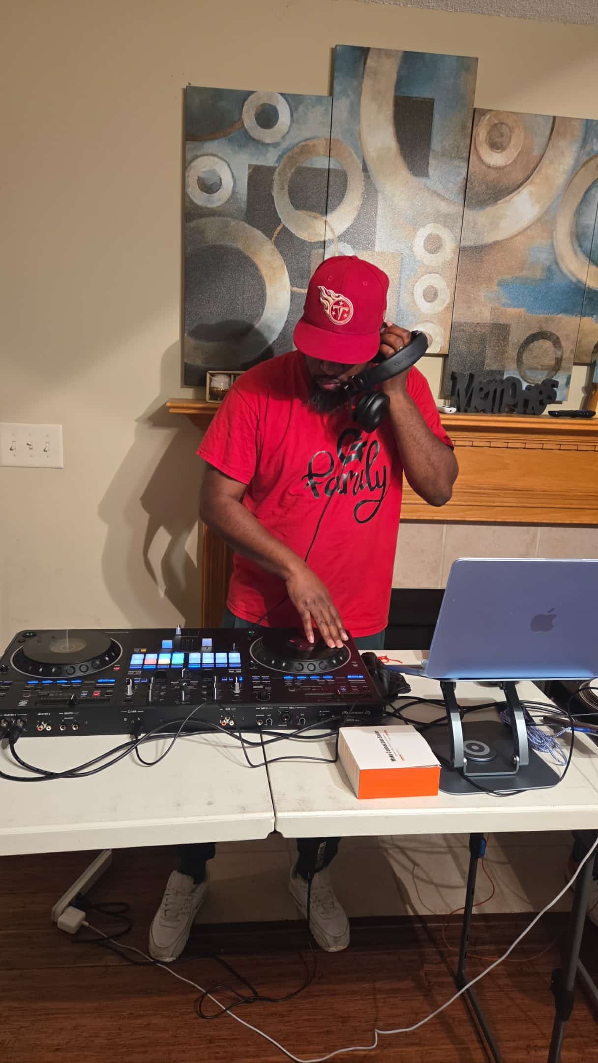 Person wearing a red cap and shirt, DJ equipment on table, abstract artwork in background.