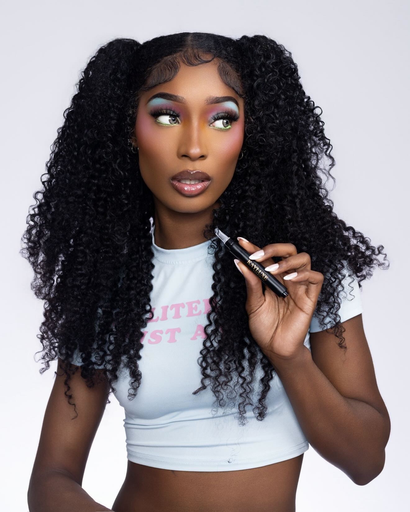 Woman with curly hair holding a makeup brush, wearing a white crop top, looking to the side, against a plain background.