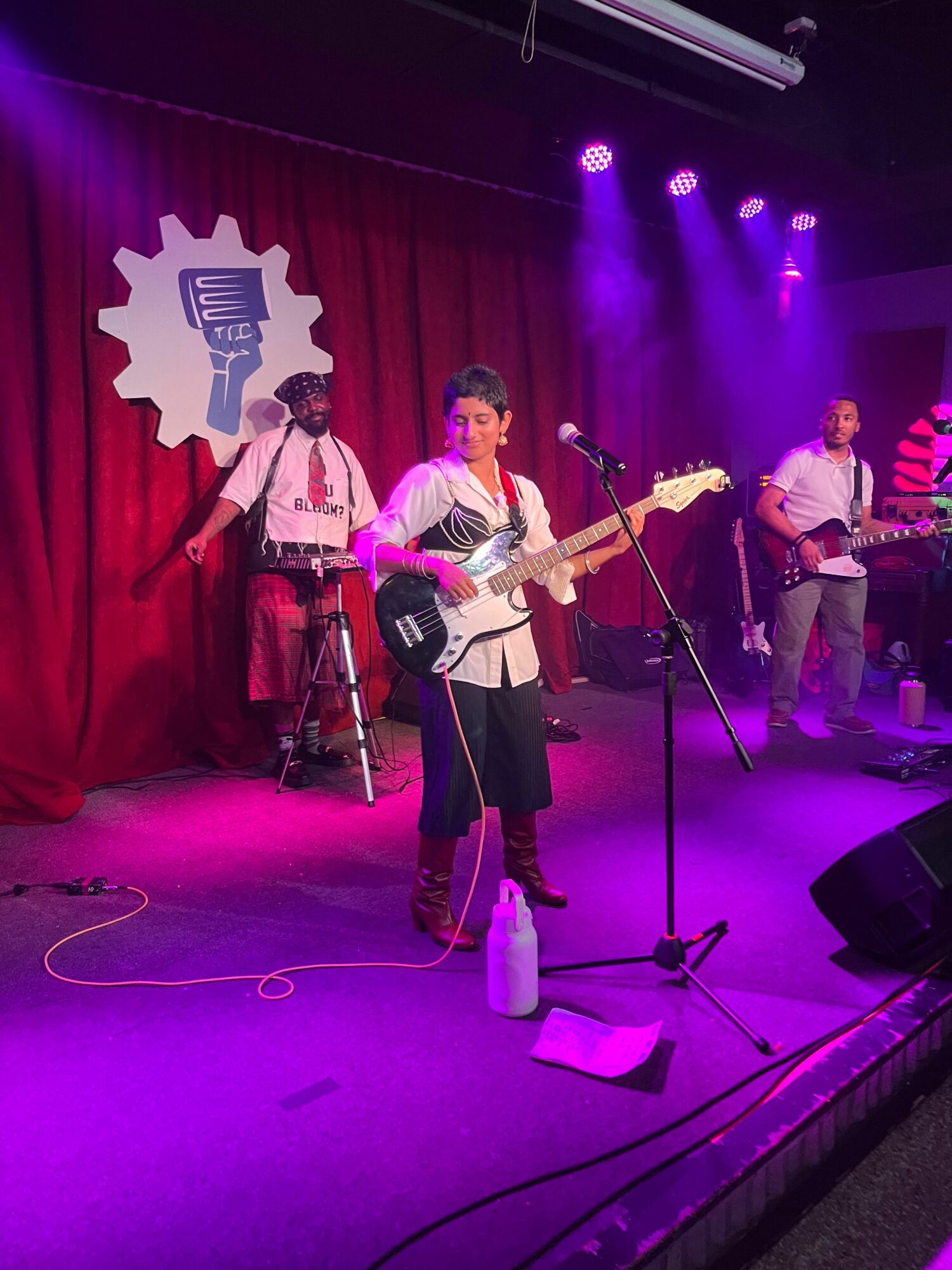 Band performing on stage with red curtain background, purple stage lighting, and musical instruments, including guitars and drums.