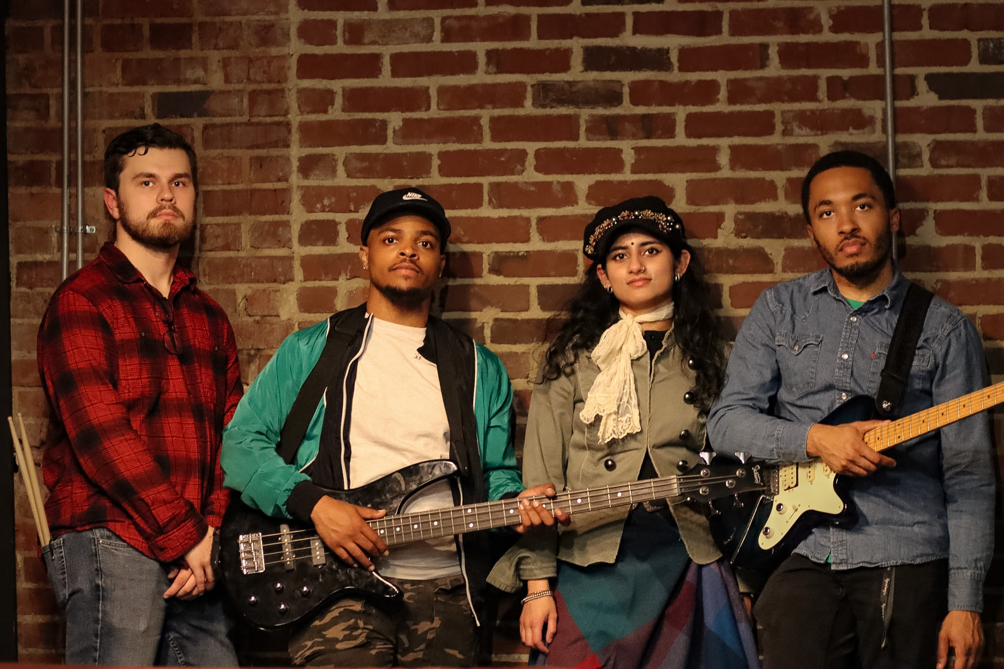 Four people standing against a brick wall, two holding guitars, one wearing a cap, and one in a red plaid shirt.