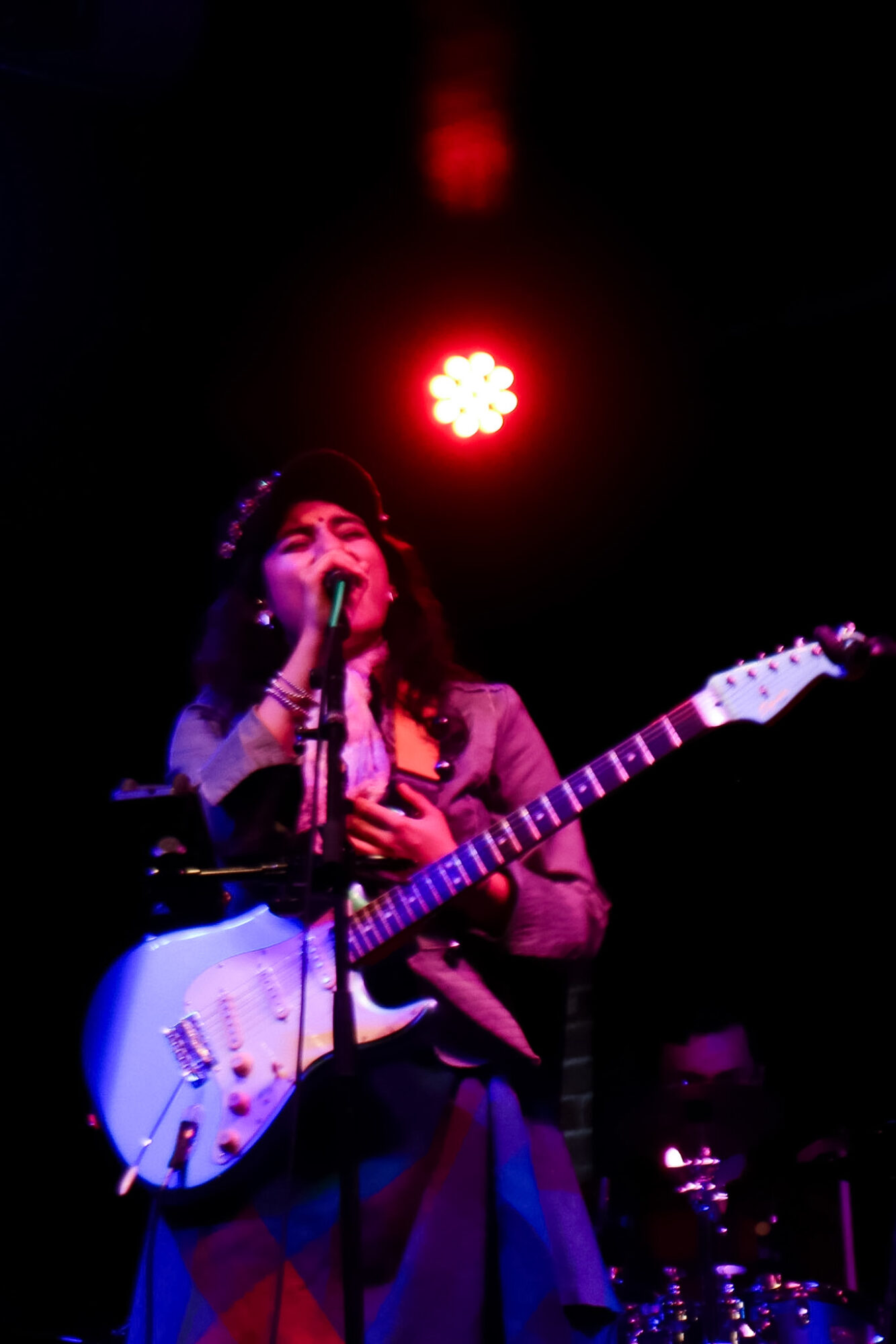 Person singing into microphone while holding an electric guitar on stage with red lighting above