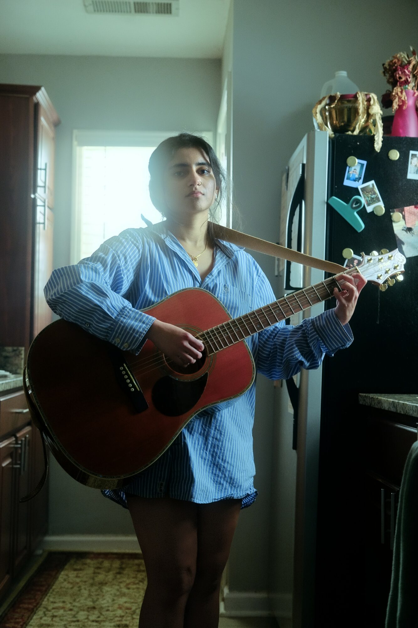 Young woman with dark hair in a ponytail playing an acoustic guitar indoors, wearing a blue shirt, standing near a refrigerator.