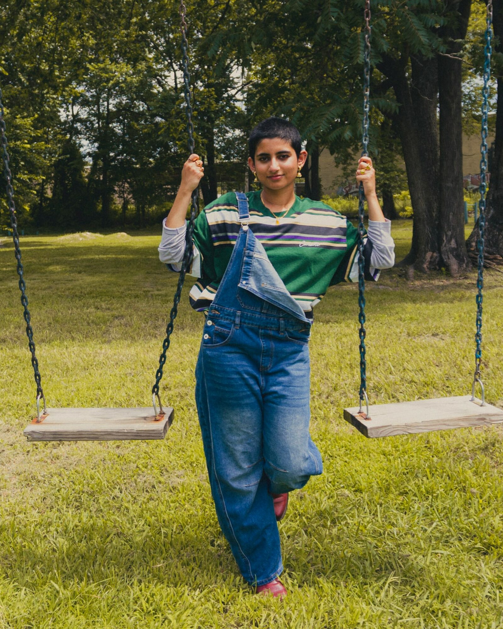 Young woman standing on grass between two empty swings in a park, holding swing chains, trees in background.