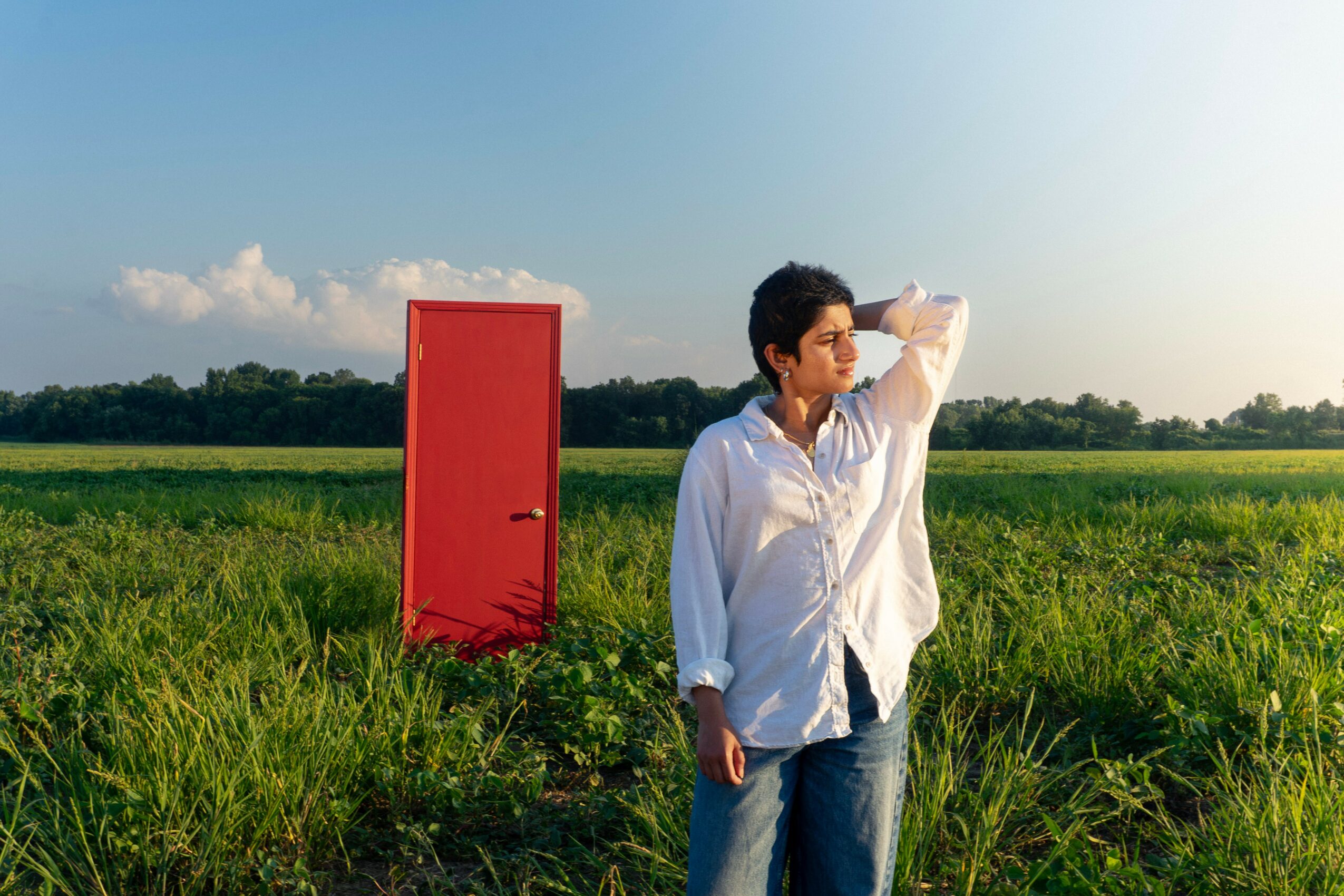 A person stands in a grassy field with a red door behind them, looking into the distance on a sunny day.