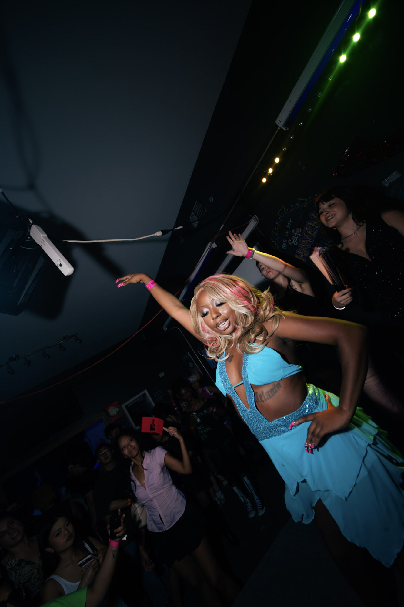 A woman in a blue costume with blonde hair dances at a party, surrounded by people in a dark room with colorful lights.
