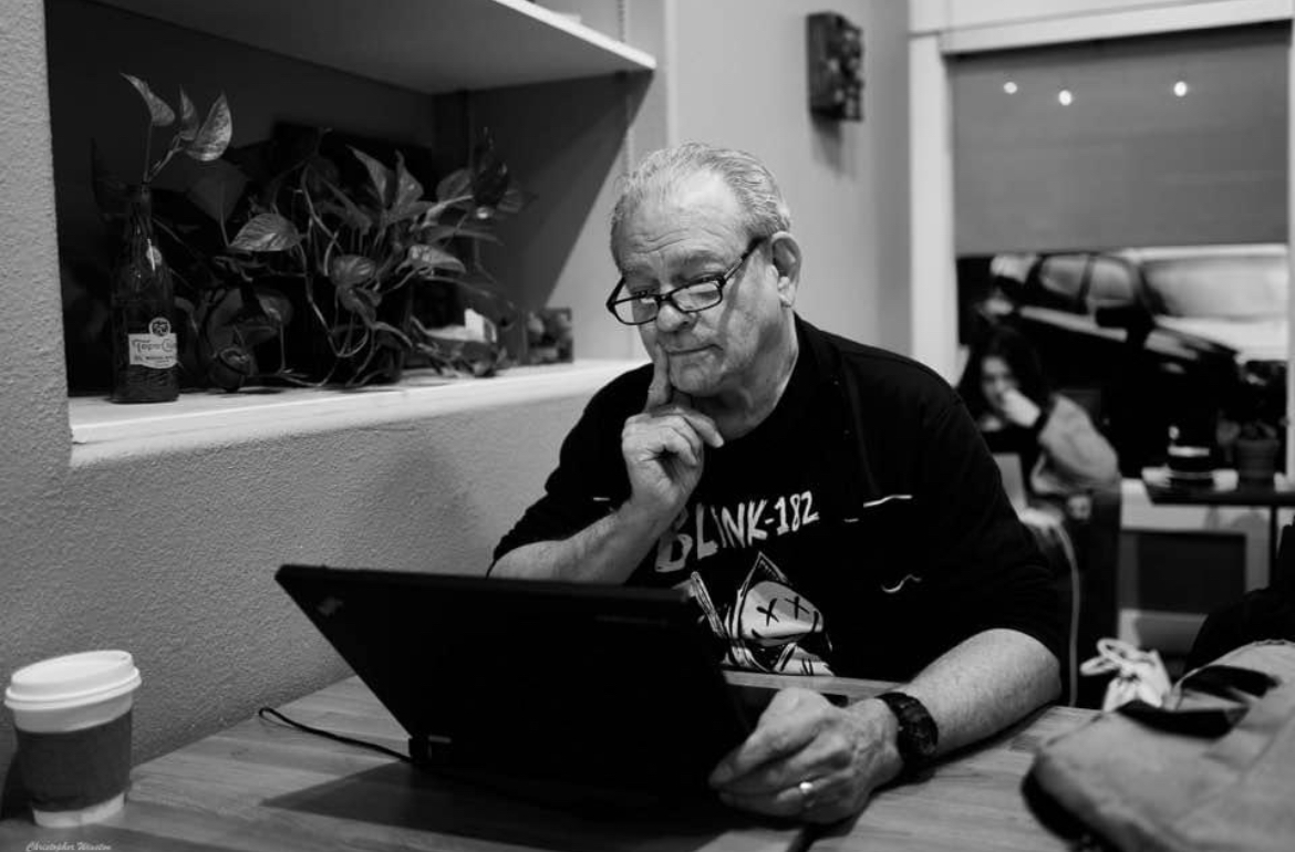 Man with glasses working on a laptop at a table in a cafe, with plants and a window in the background.