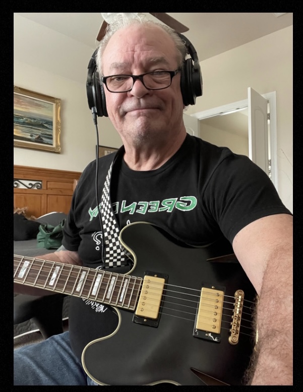 Older man with glasses and headphones holding a black electric guitar, smiling, in a room with a painting and open door.