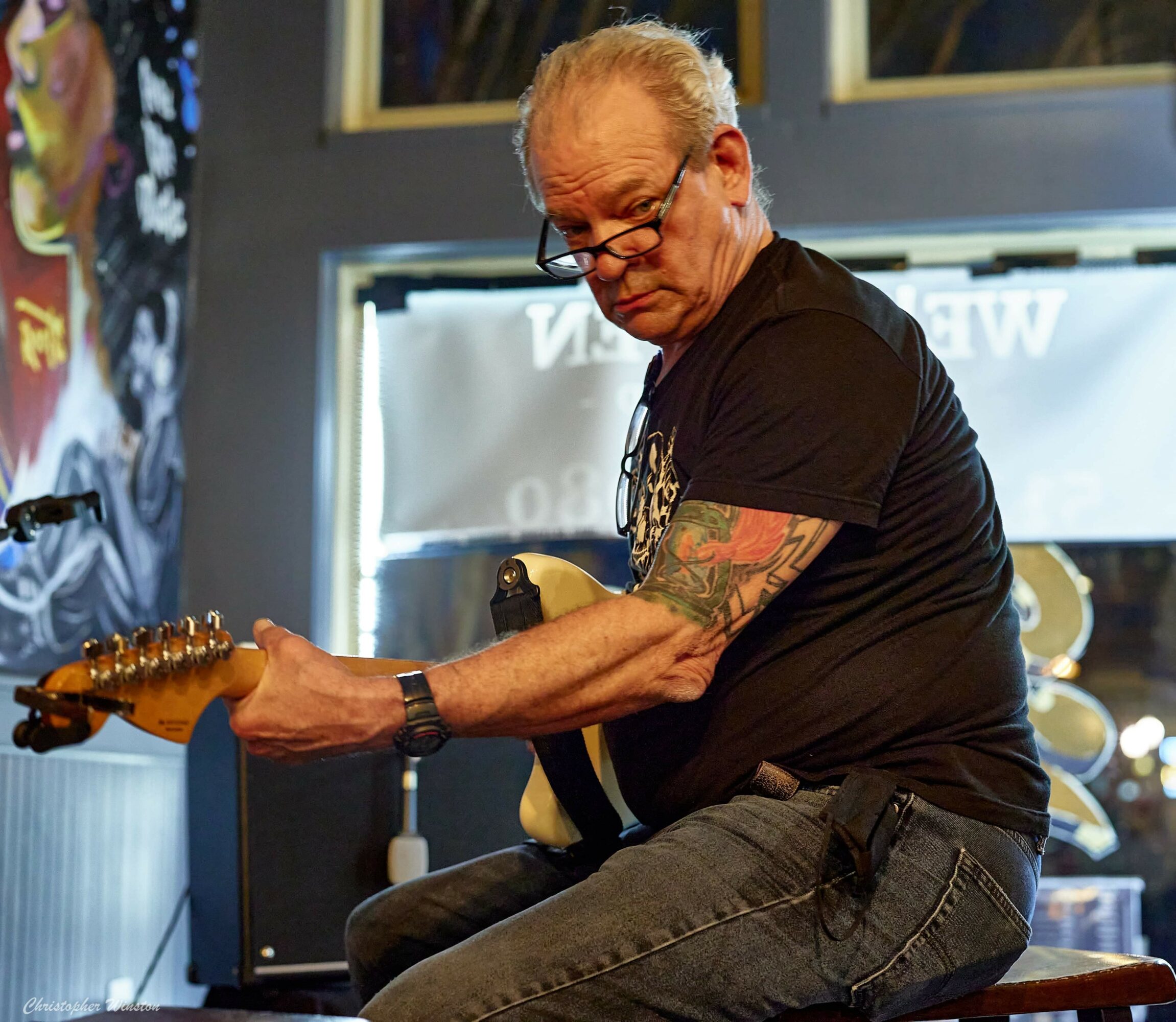 Man with glasses and tattoos playing an electric guitar on stage, focused on his instrument.