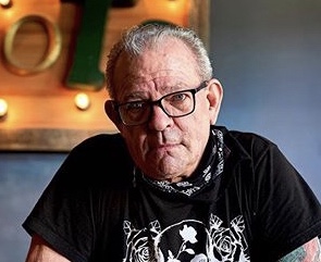 Older man with glasses and short hair, wearing a black T-shirt and a patterned bandana around his neck, sitting indoors.