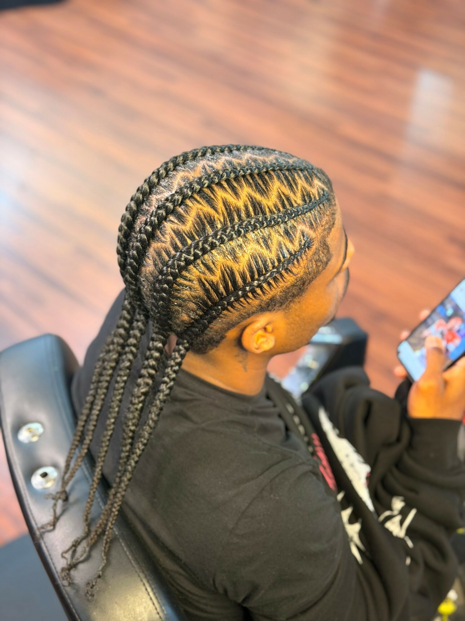 Person with braided hairstyle sitting in a chair, looking at a phone, with a wooden floor background.