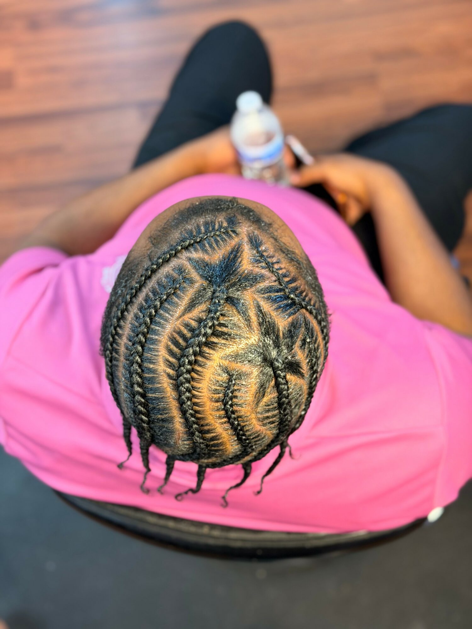 Top view of a person with braided hair wearing a pink shirt, holding a water bottle, sitting on a wooden floor.