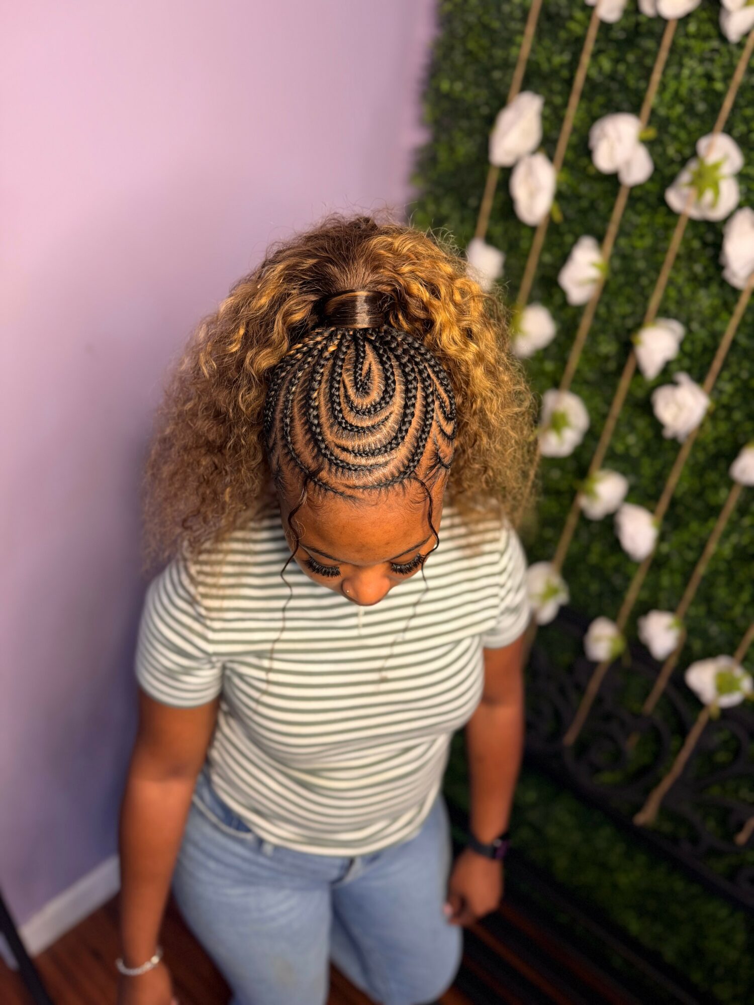 Top view of a woman with curly hair and intricate braids, wearing a striped shirt, standing near a floral wall.