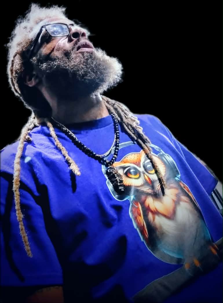 Person with glasses and dreadlocks wearing a blue shirt with a cat graphic, looking upward against a black background.