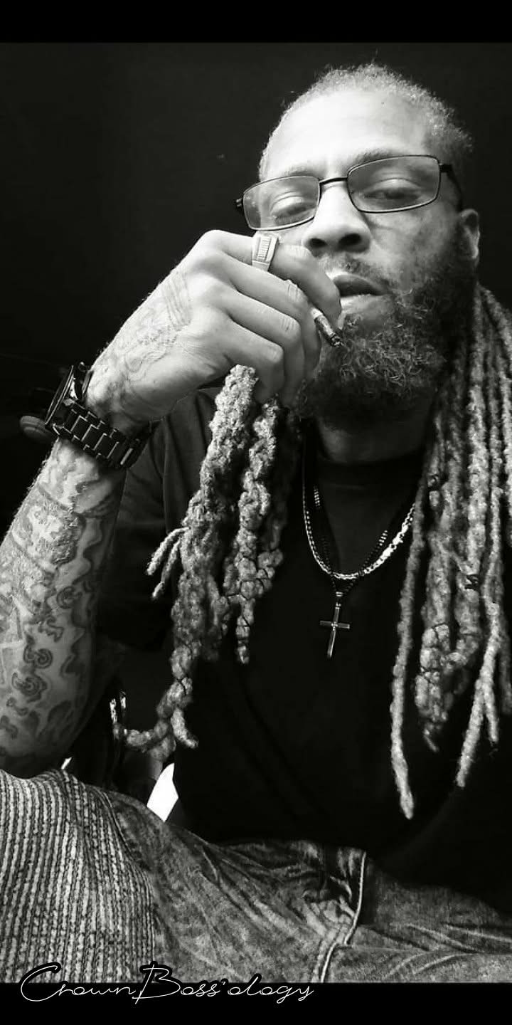 Man with glasses and dreadlocks, resting his chin on his hand, wearing a watch and chain, in black and white.