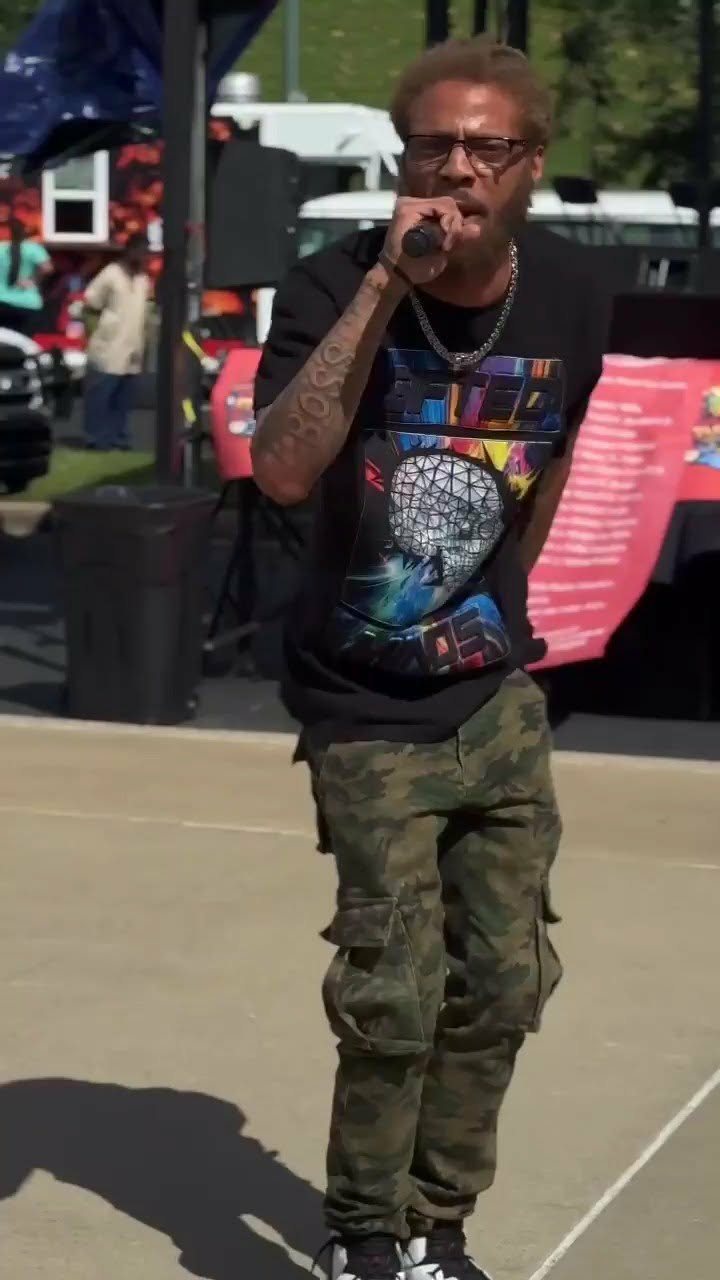 Man with glasses and tattoos singing into a microphone outdoors, wearing a graphic t-shirt and camouflage pants.